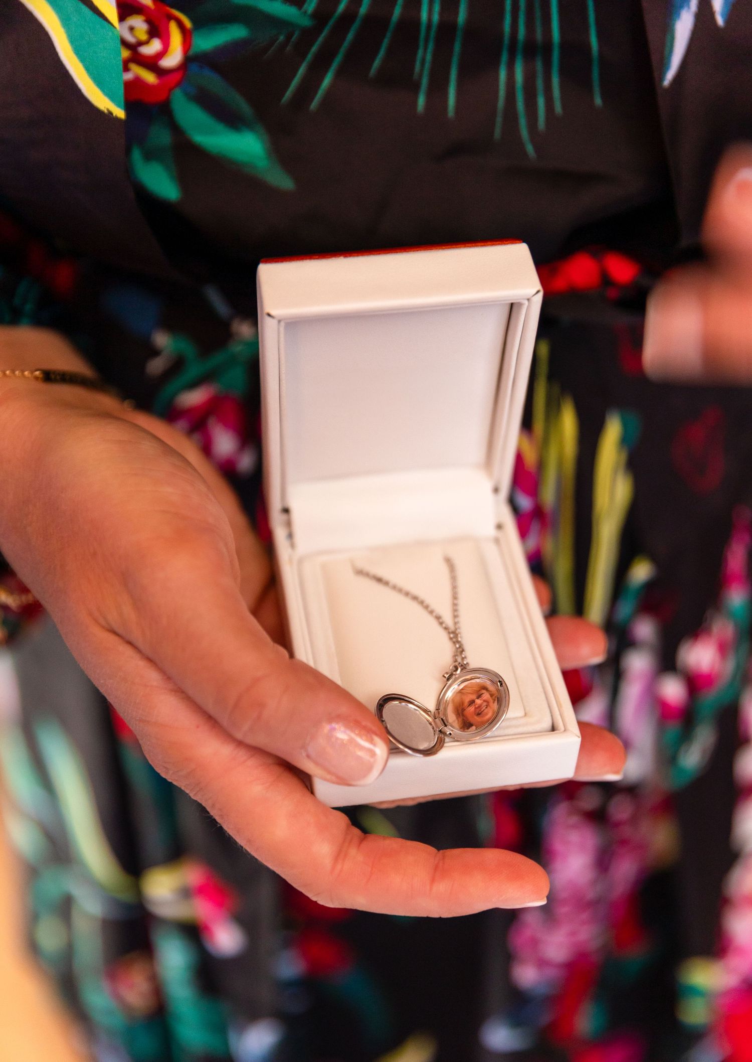 Hand holding an open white jewelry box displaying a necklace with a pendant against a floral patterned background.