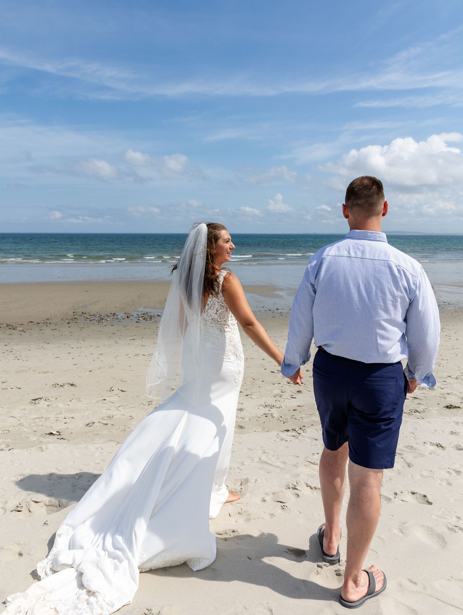A couple in wedding attire walks along a pristine beach with white sand and blue ocean on a sunny day.
