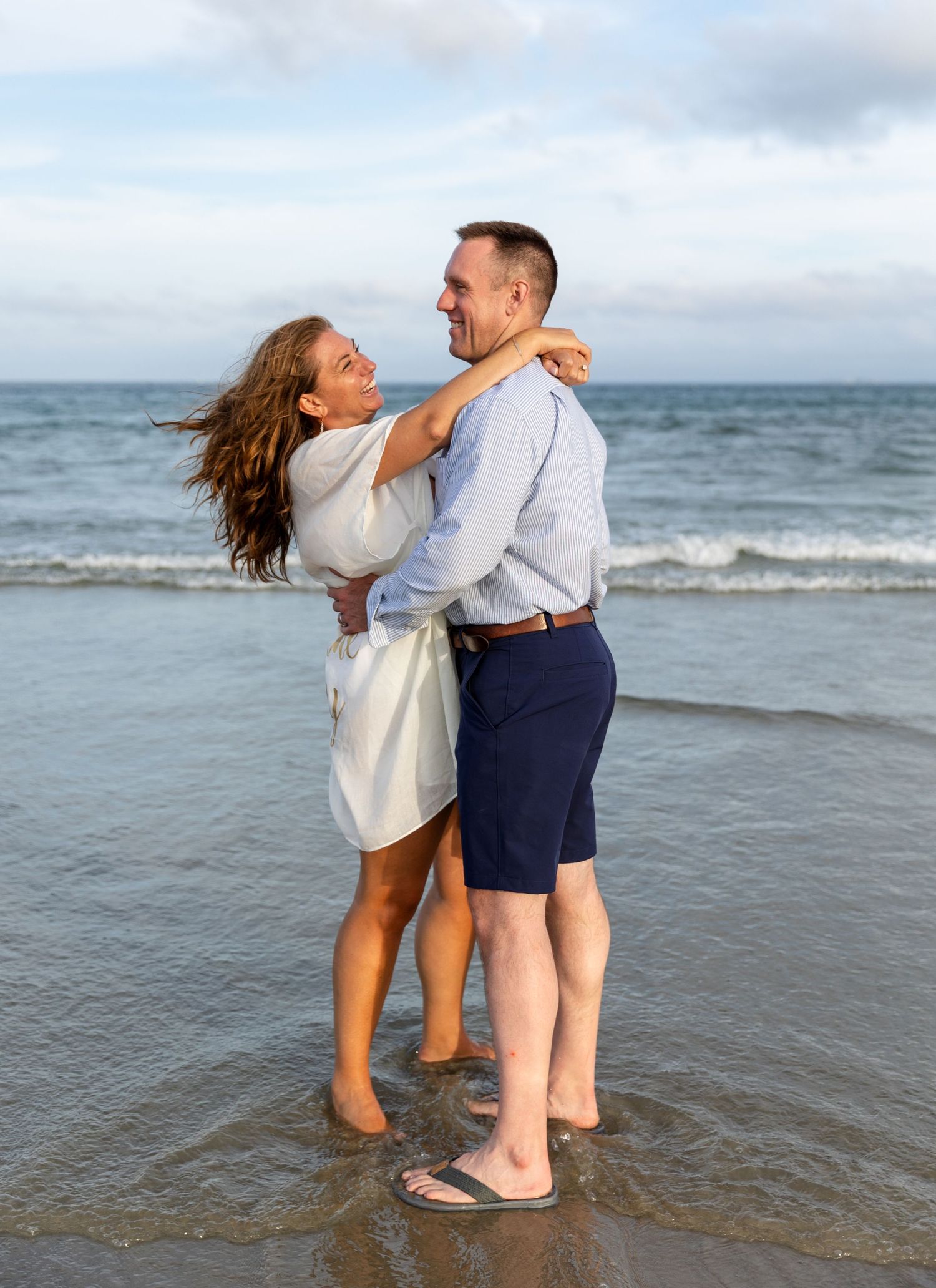 Two playful people embrace while standing in the ocean waves at the beach shore enjoying a joyful candid moment together.