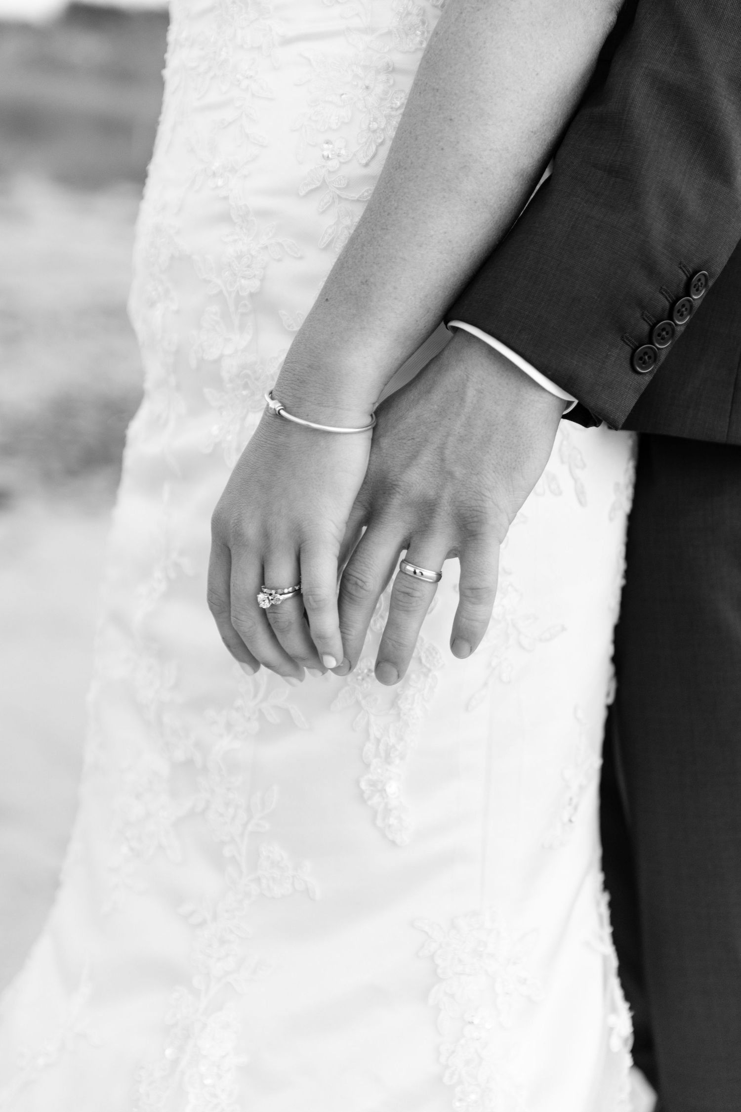 Black and white close-up photo of newlywed couple's hands with wedding rings and white lace dress detail.