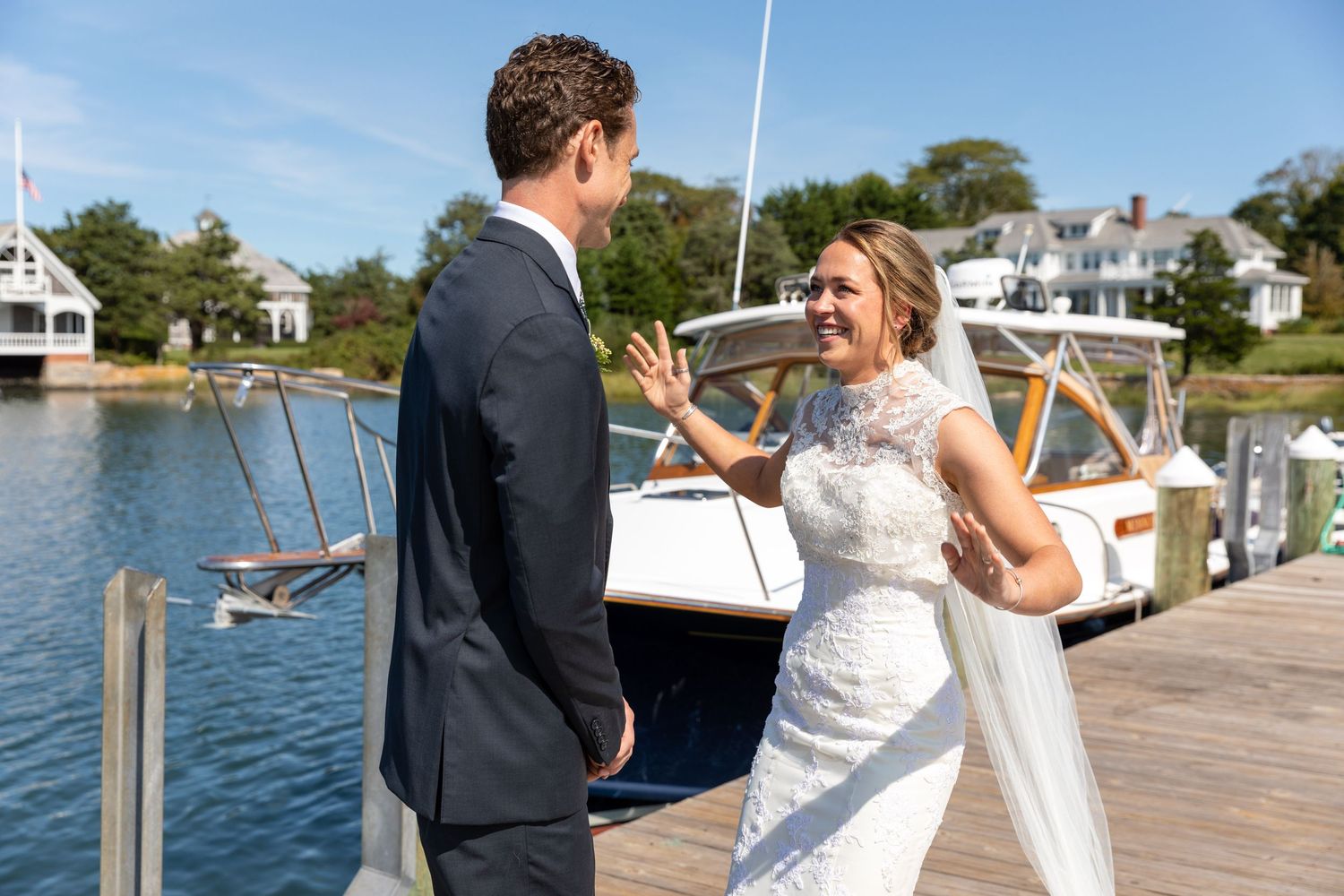Bride and groom share a joyful moment together on a wooden dock with boats and waterfront homes in background.