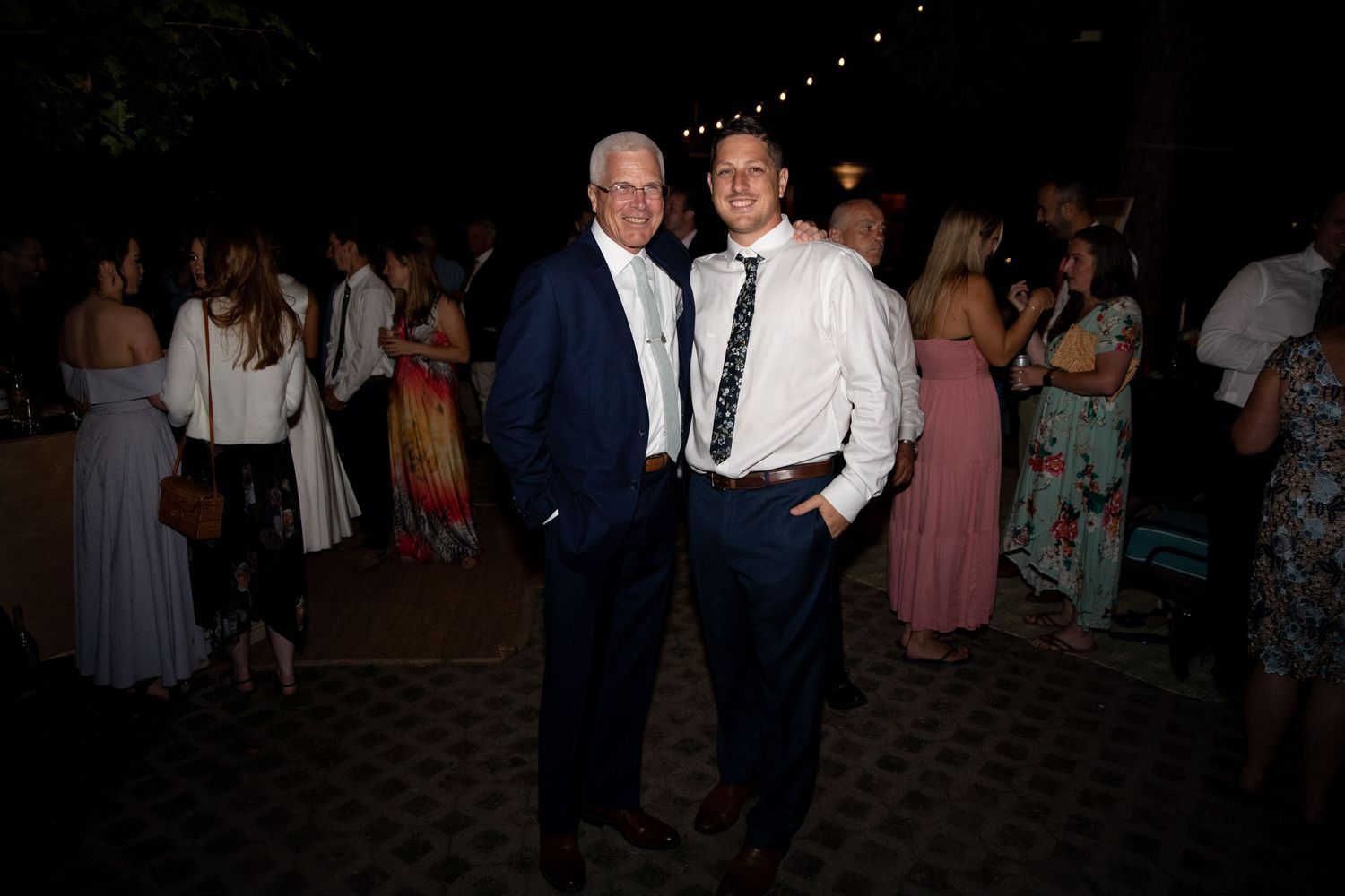 Two people in formal attire stand together at an evening event with other guests visible in the background.