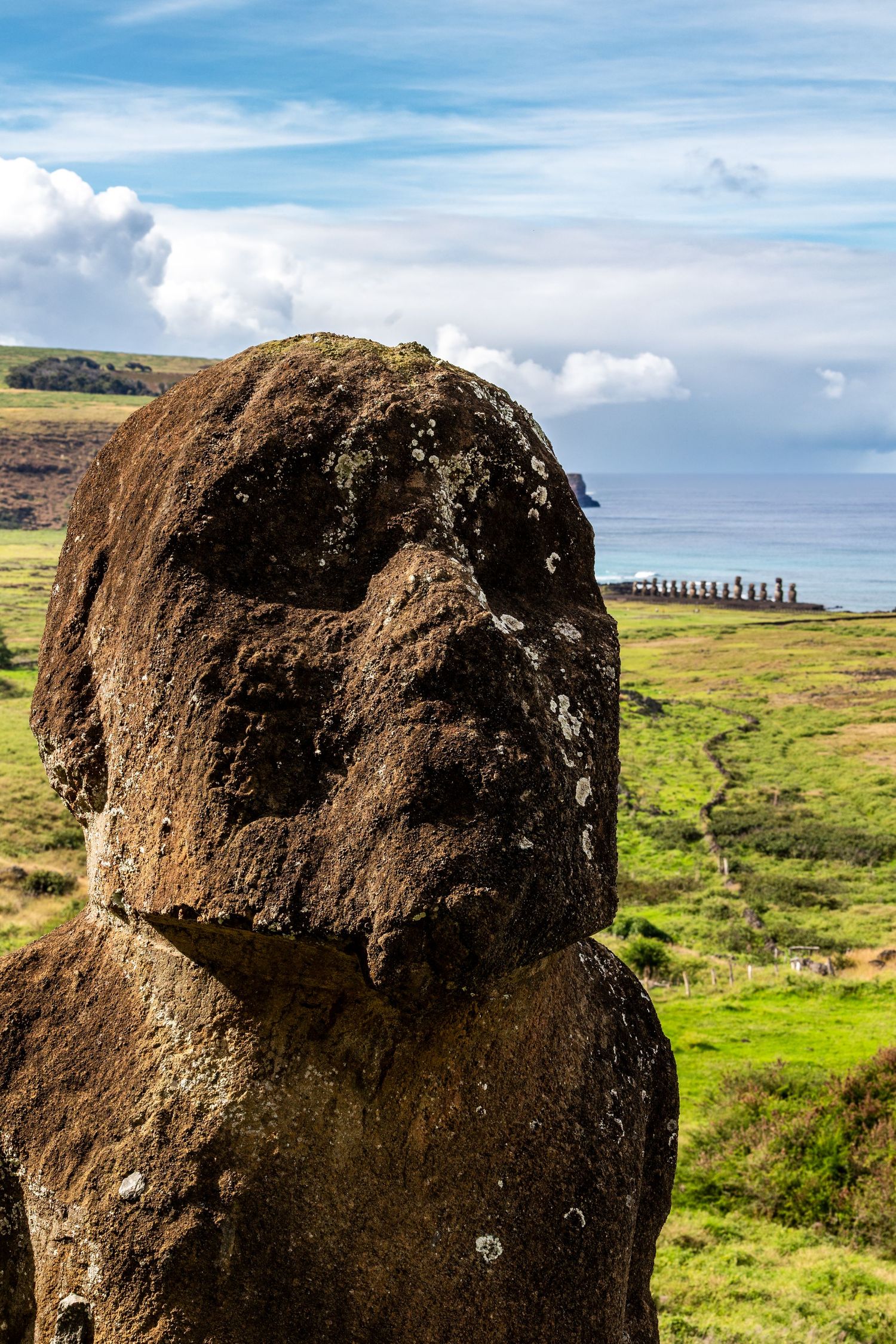 Giant stone moai heads rise from the grassy landscape of Easter Island against a bright blue sky with white clouds.