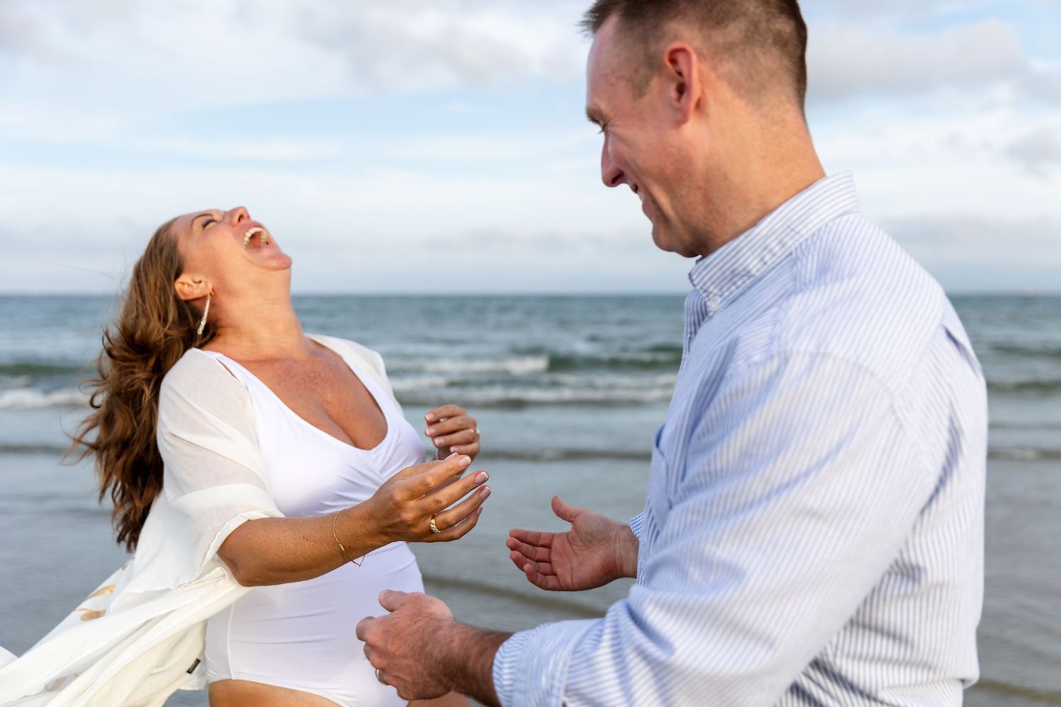 Couple laughing together on beach at sunset in casual white attire.