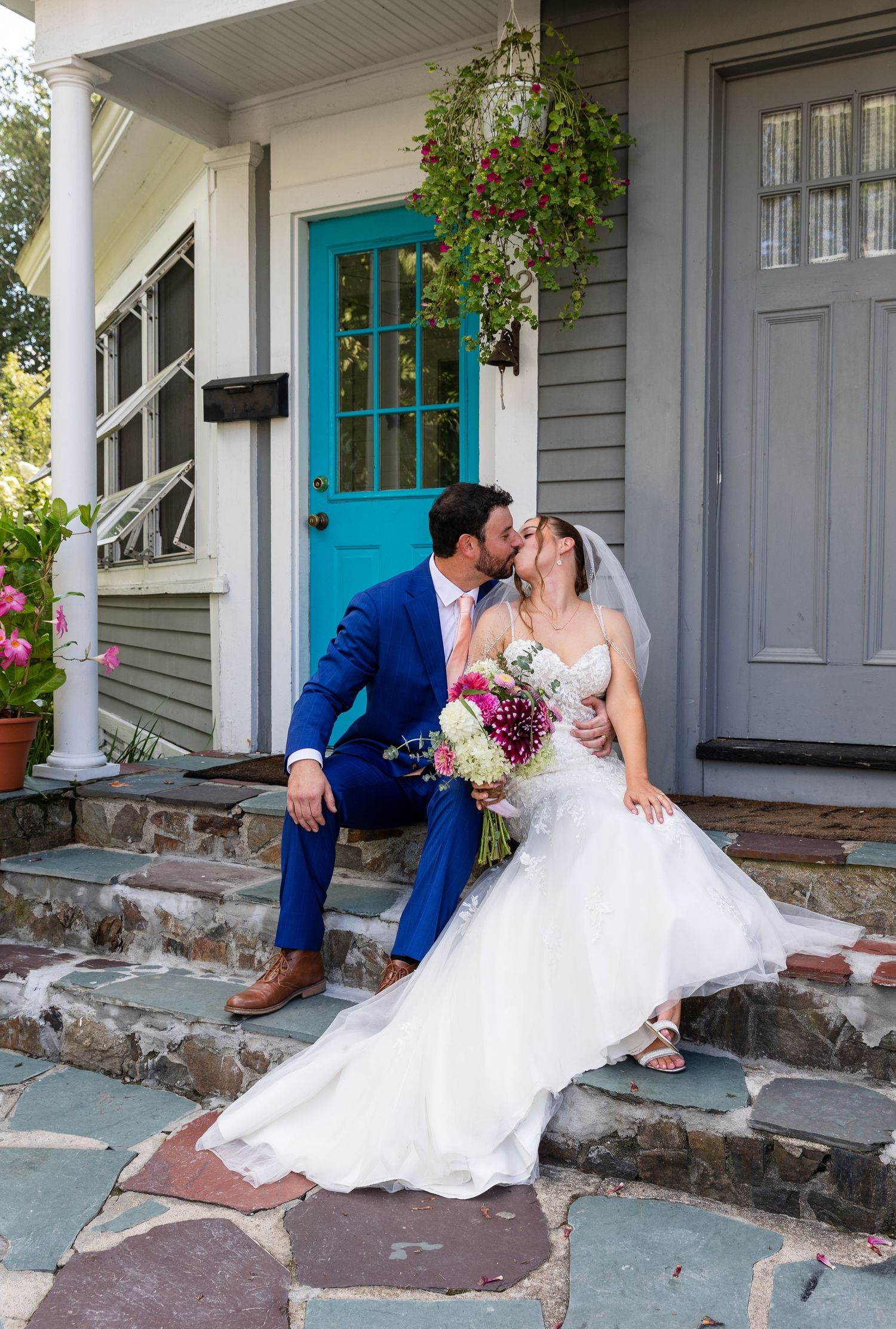 Newlyweds share a romantic moment on stone steps outside house with bright turquoise door and hanging floral baskets.
