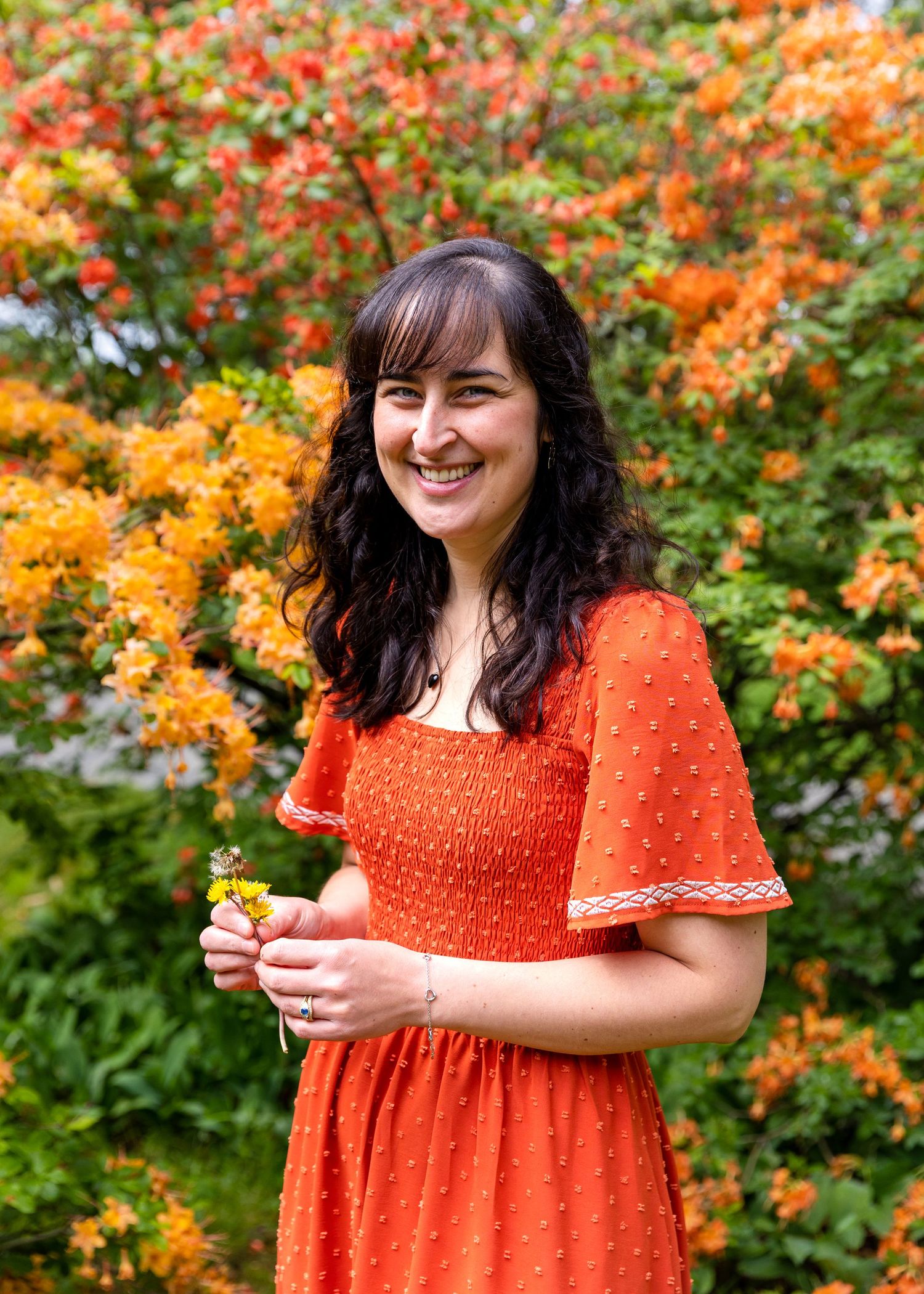 Someone in a bright orange smocked dress stands smiling in front of vibrant fall foliage with orange and red colors.