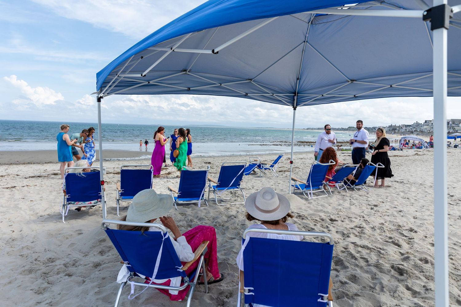 Beach gathering under a blue canopy tent with folding chairs set up on the sand during a sunny day at the seaside.