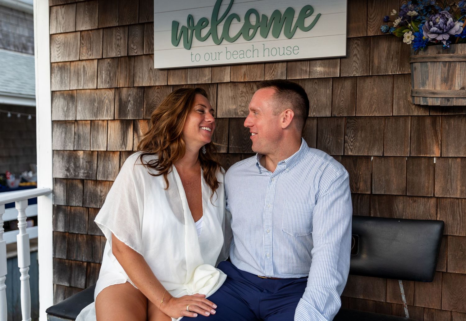 Two people share a laugh while sitting on a porch bench beneath a rustic welcome sign on a cedar-shingled exterior.