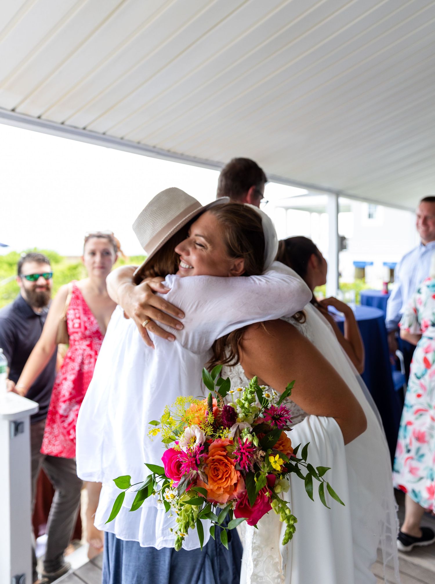 Two people share a warm embrace at an outdoor gathering while holding a vibrant bouquet of pink and orange flowers.