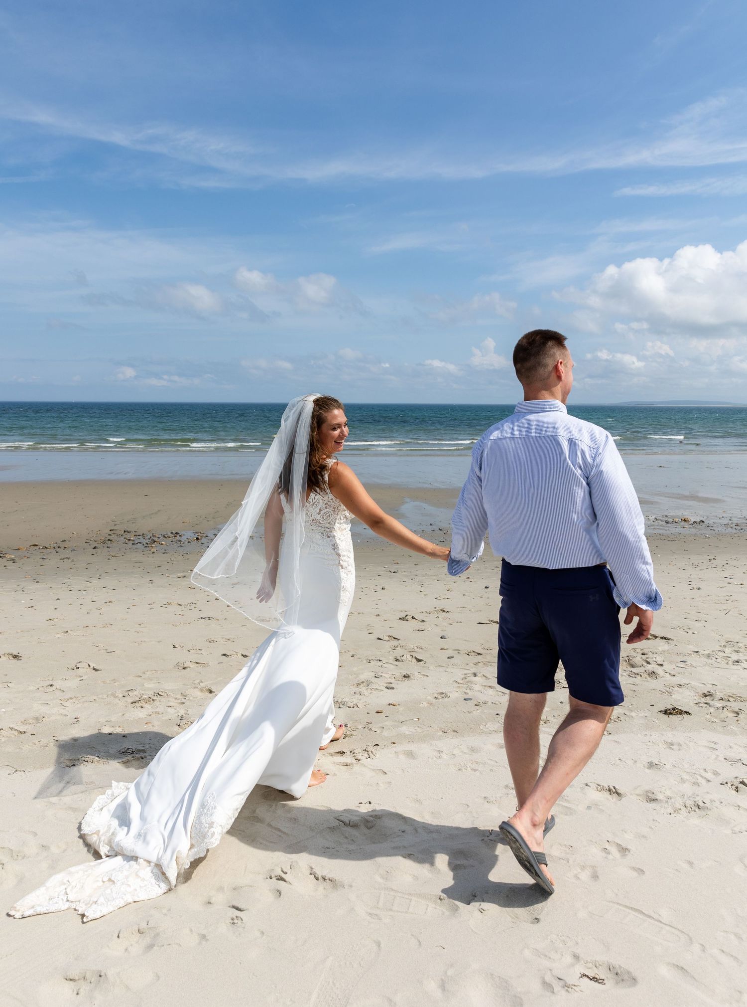 Couple walking hand in hand on sunny beach as bride's white wedding dress and veil flow in ocean breeze.