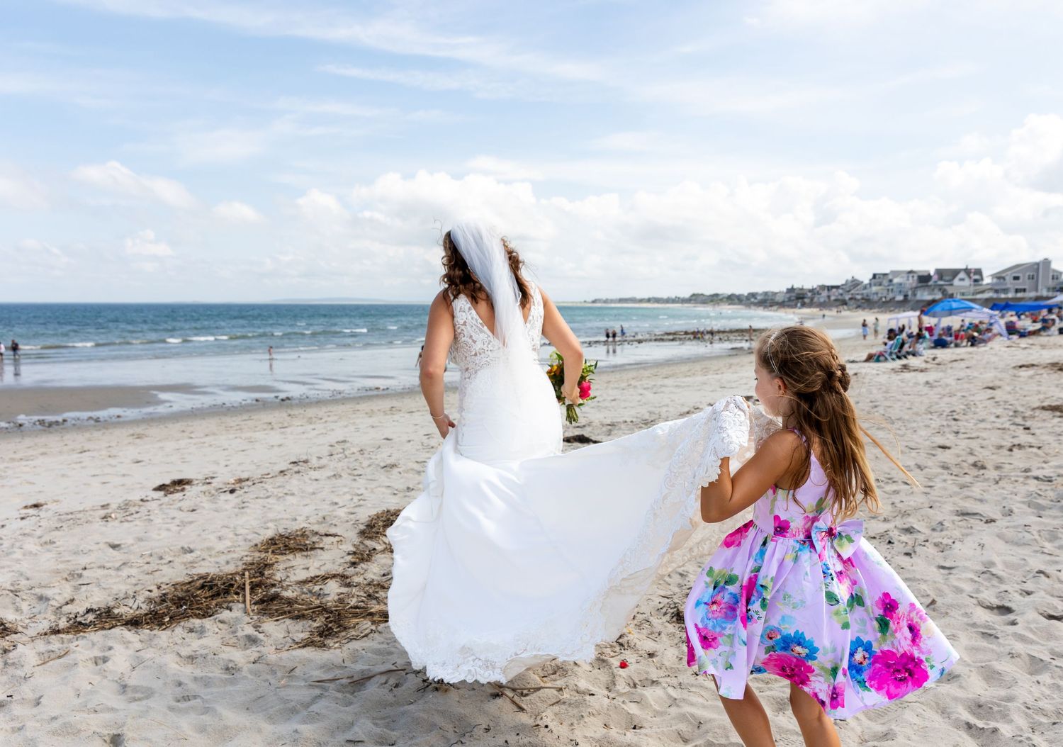 Two people dance and twirl joyfully on a sunny beach with the ocean and beachgoers visible in the background.