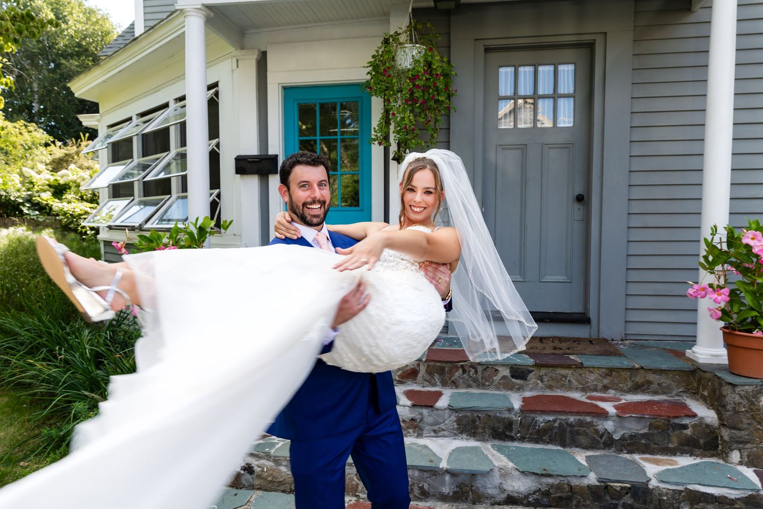 A joyful couple celebrates their wedding day in front of a gray house with a turquoise door.