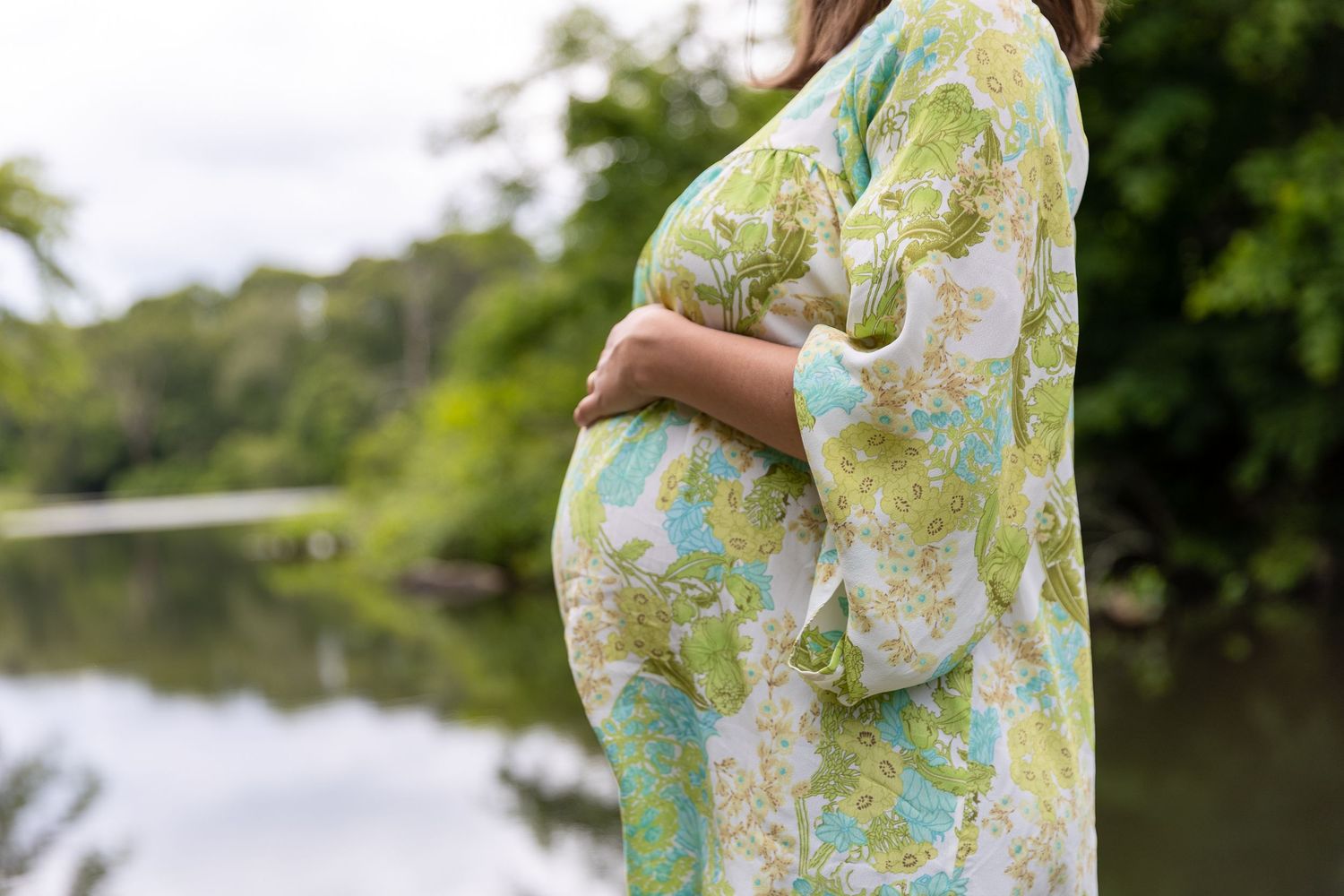 A pregnant person wearing a floral kimono stands beside a peaceful lake while holding their baby bump.