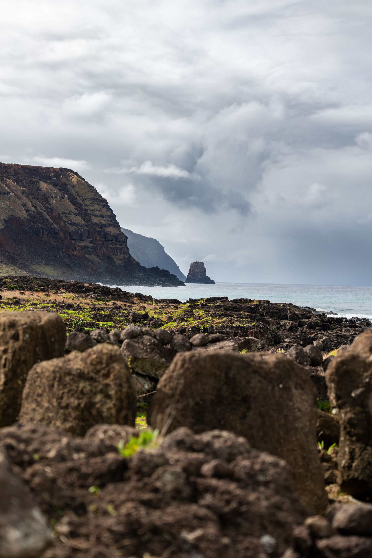 Dramatic coastal landscape with rocky cliffs and sea stacks along a misty coastline viewed through volcanic rocks in foreground.