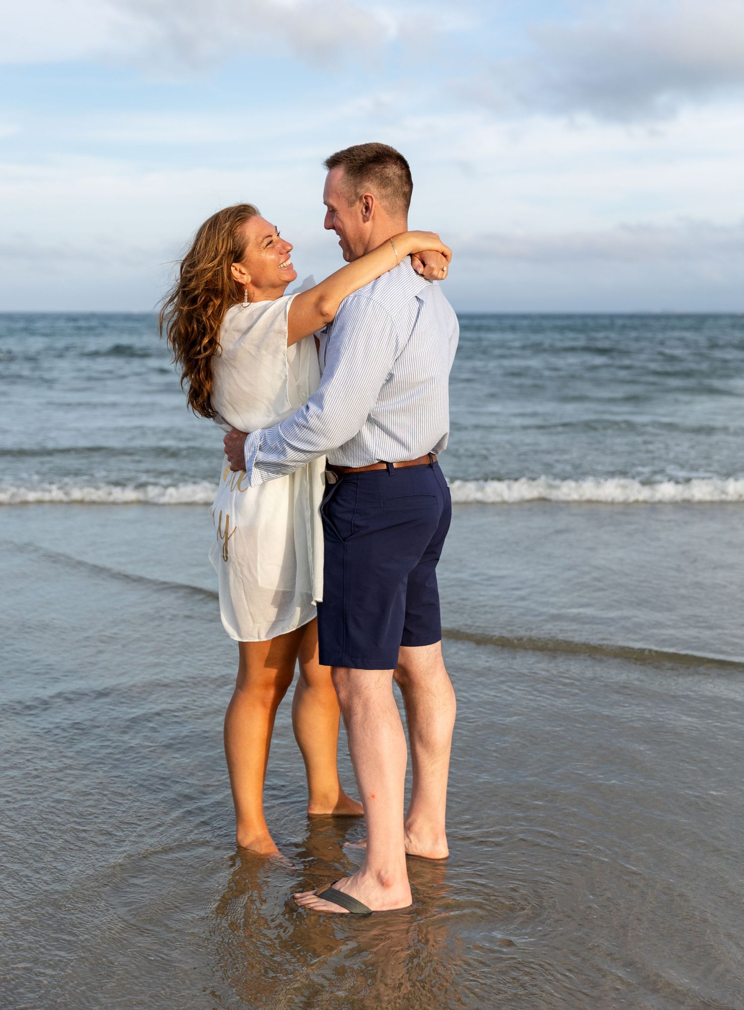 A couple embraces while standing barefoot in shallow ocean waves at the beach during a cloudy day.