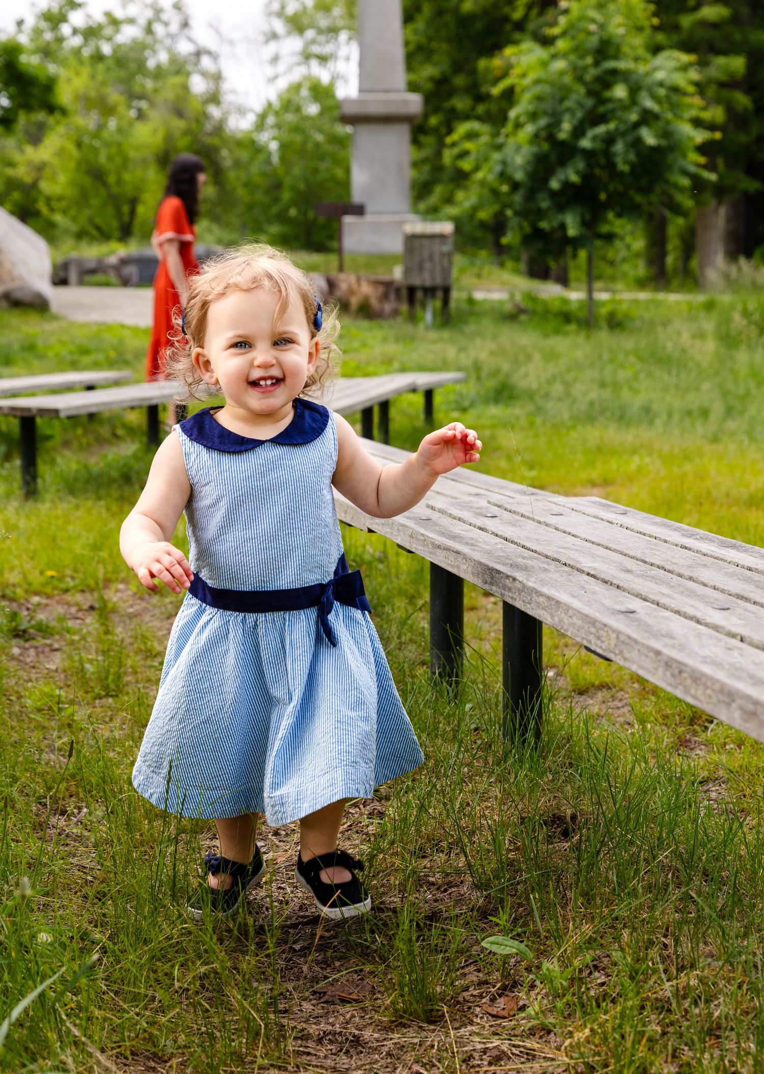 A small child wearing a light blue dress and sandals happily runs along a wooden bench in a garden setting.