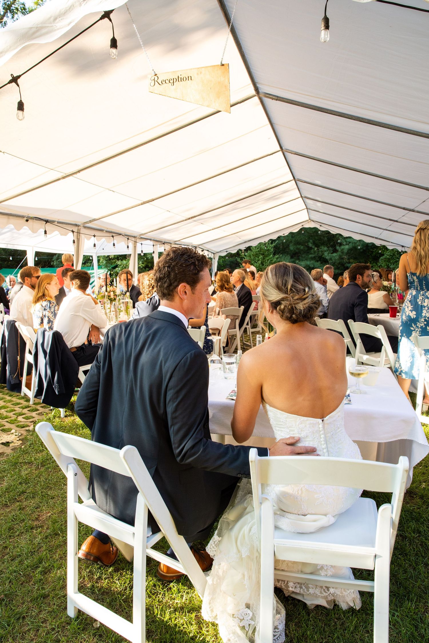 Wedding reception guests seated under an outdoor tent with string lights and white folding chairs during a summer celebration.