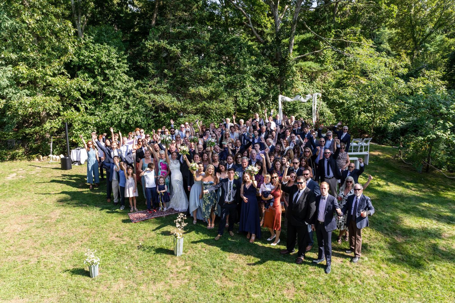 Large group of formally dressed people gathered outdoors for a group photo in a sunny garden setting.