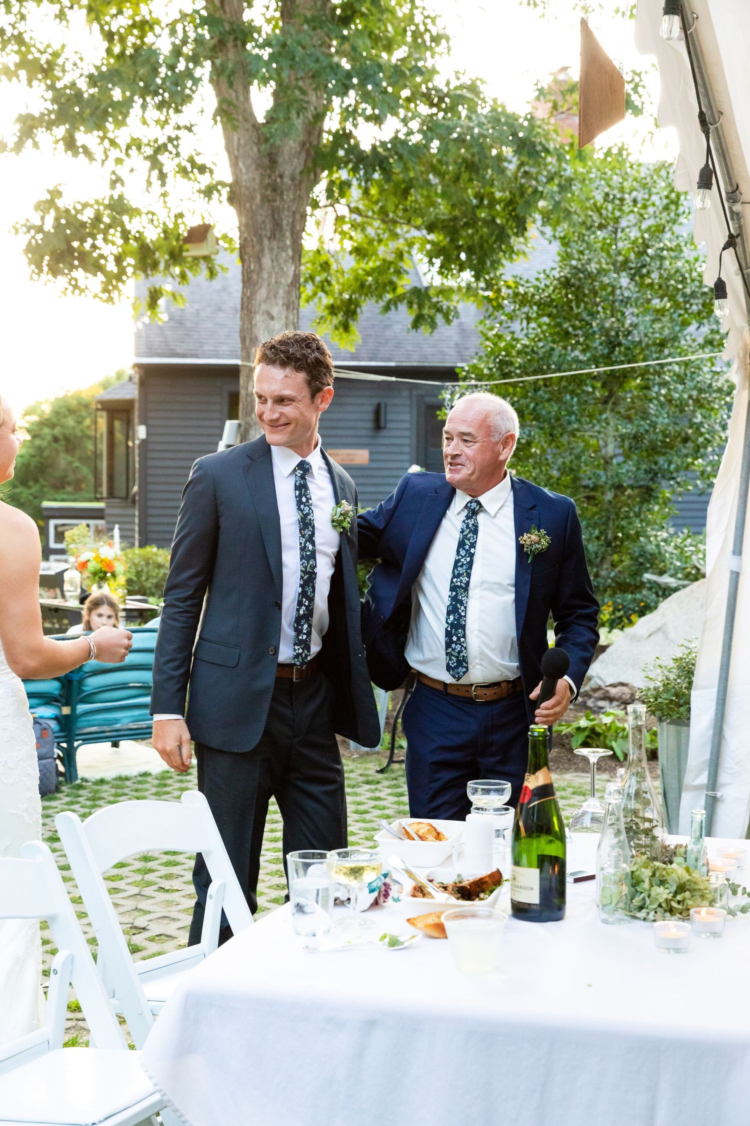Wedding party in dark suits gather around an outdoor table with white linens and champagne bottles at sunset.