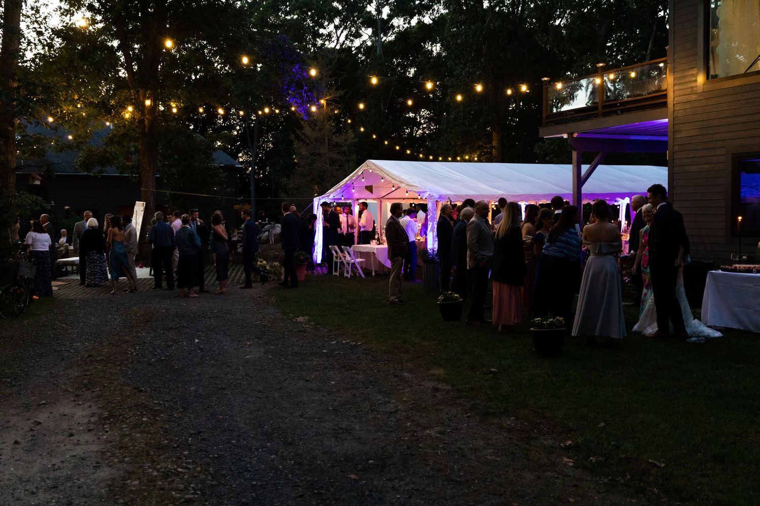 An outdoor evening party with string lights illuminating a white tent where guests gather in formal attire.