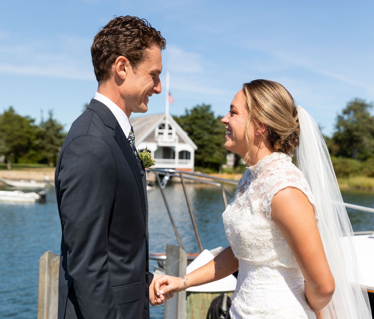 Two people in formal wedding attire share a romantic moment together on a waterfront dock during a sunny outdoor ceremony.