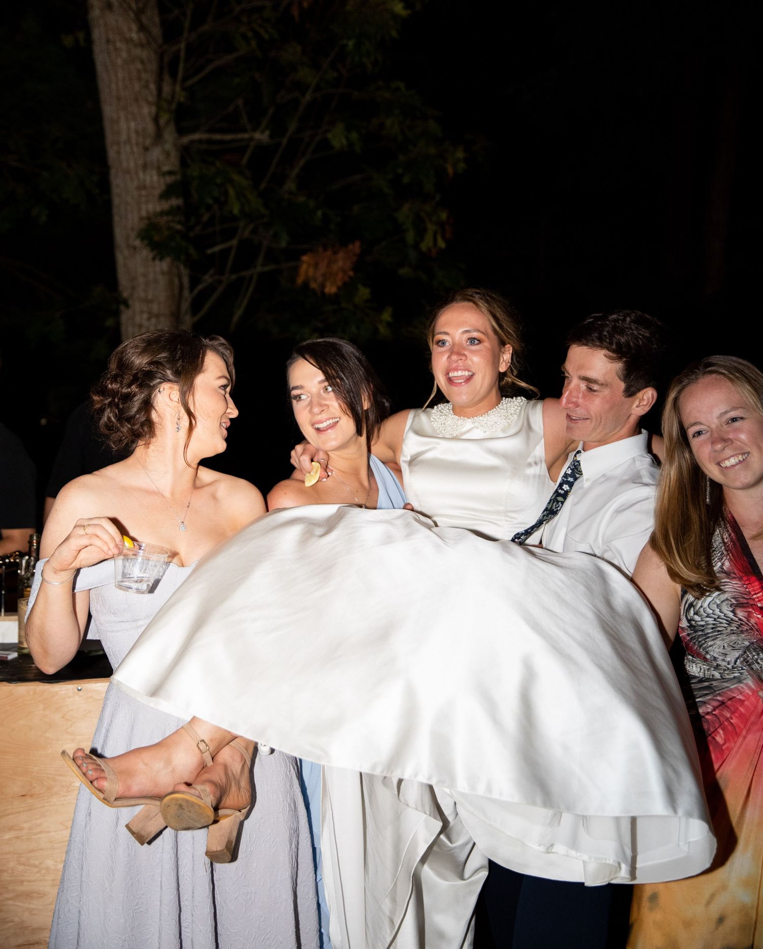 A group of wedding attendees laugh together while supporting someone in a white dress at an outdoor evening celebration.