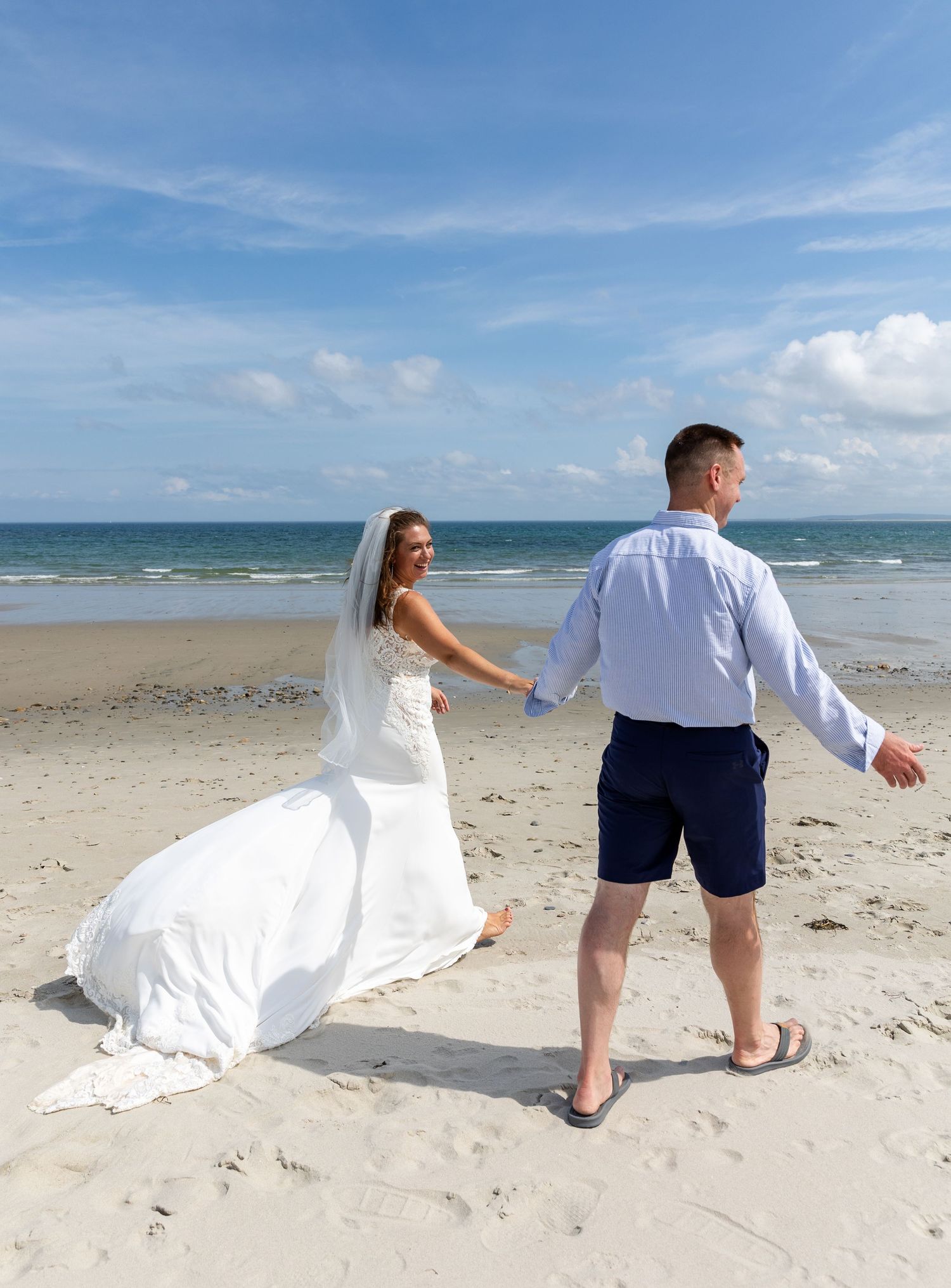 A couple in wedding attire walks on a sunny beach with blue ocean and sky in the background.