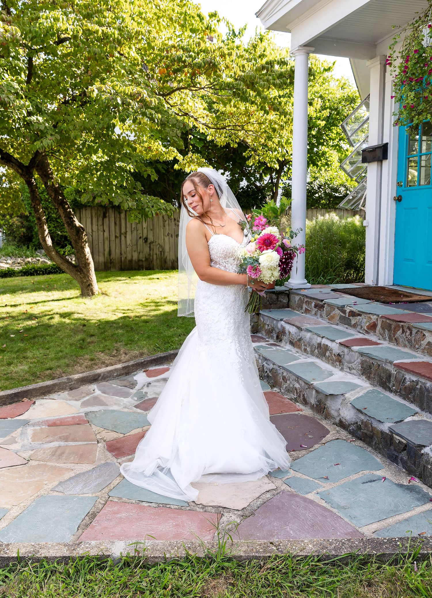 A bride stands on stone steps while holding a pink and white floral bouquet in a lush garden setting.