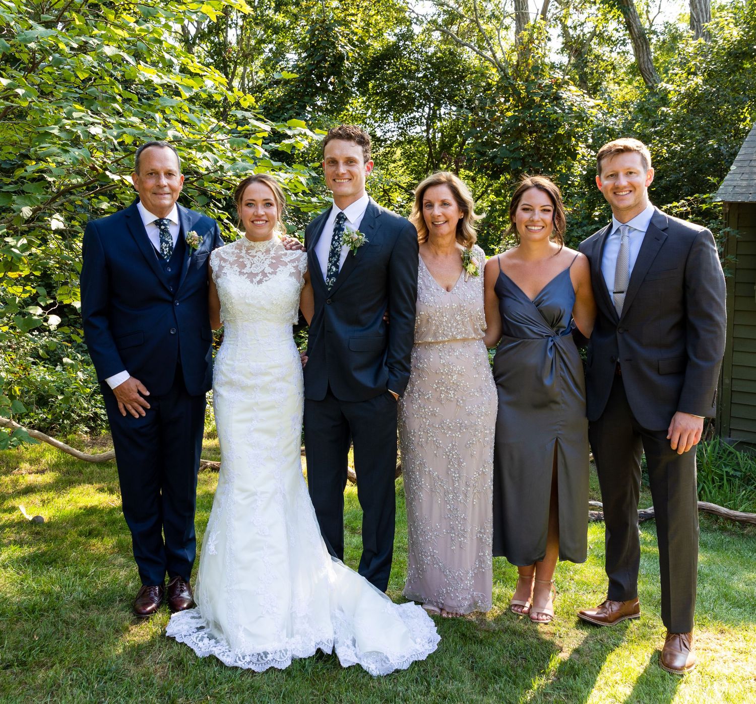 Wedding party members in formal attire pose together on sunny lawn during outdoor celebration.