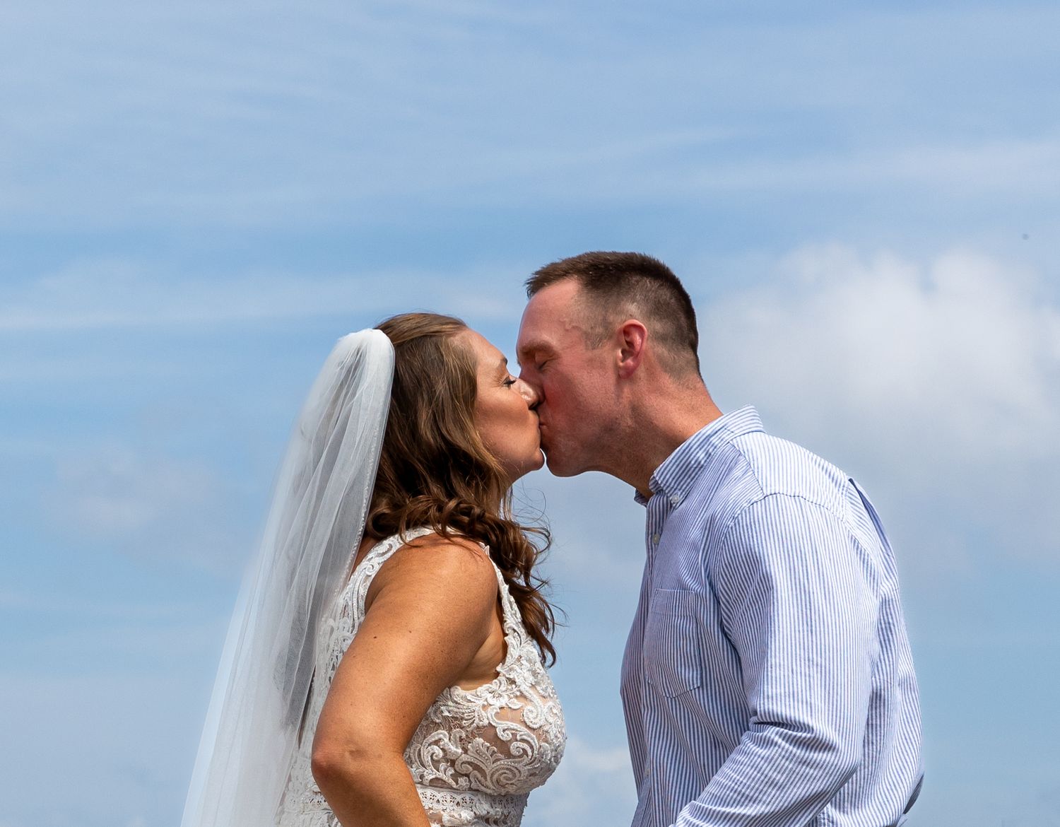 A bride and groom share a romantic kiss against a bright blue sky and white clouds on their wedding day.
