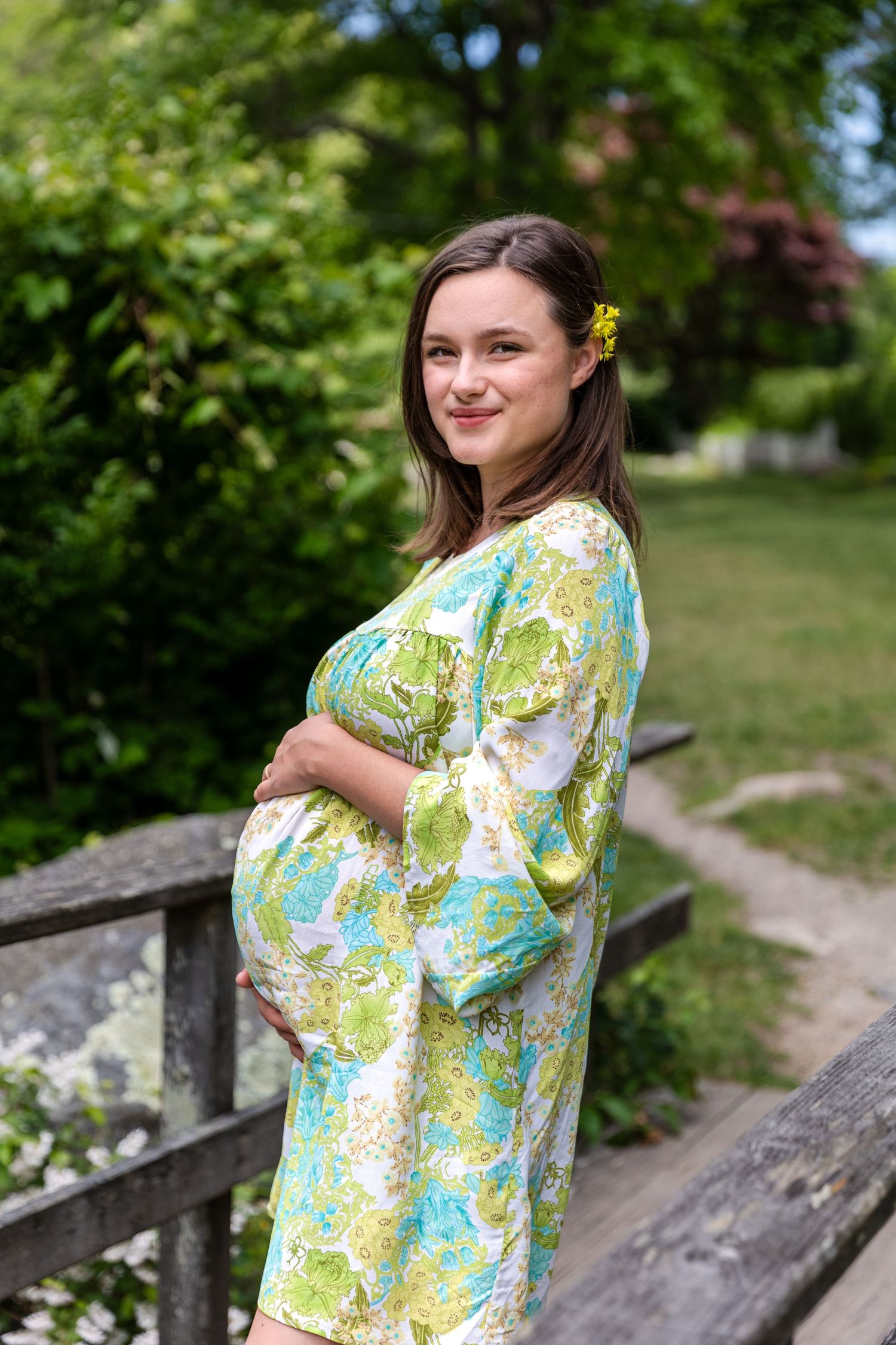 Someone wearing a colorful floral print maternity dress poses outdoors near a wooden fence surrounded by green foliage.