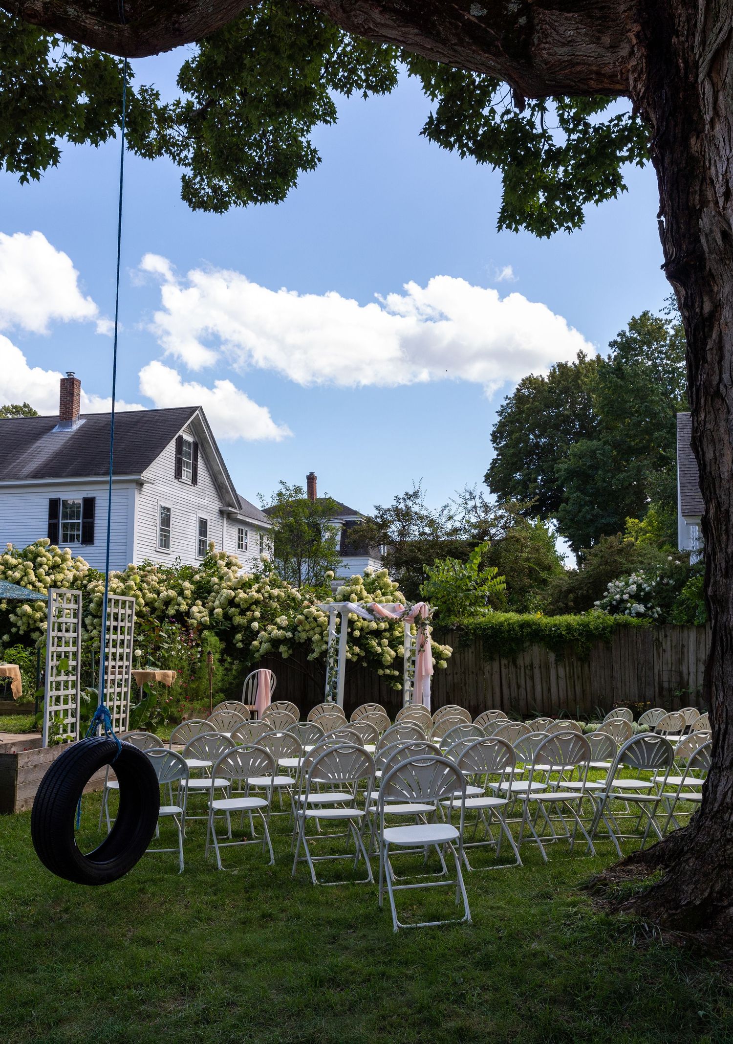 A backyard wedding setup with white folding chairs on a lawn, featuring a tire swing and white house in the background.