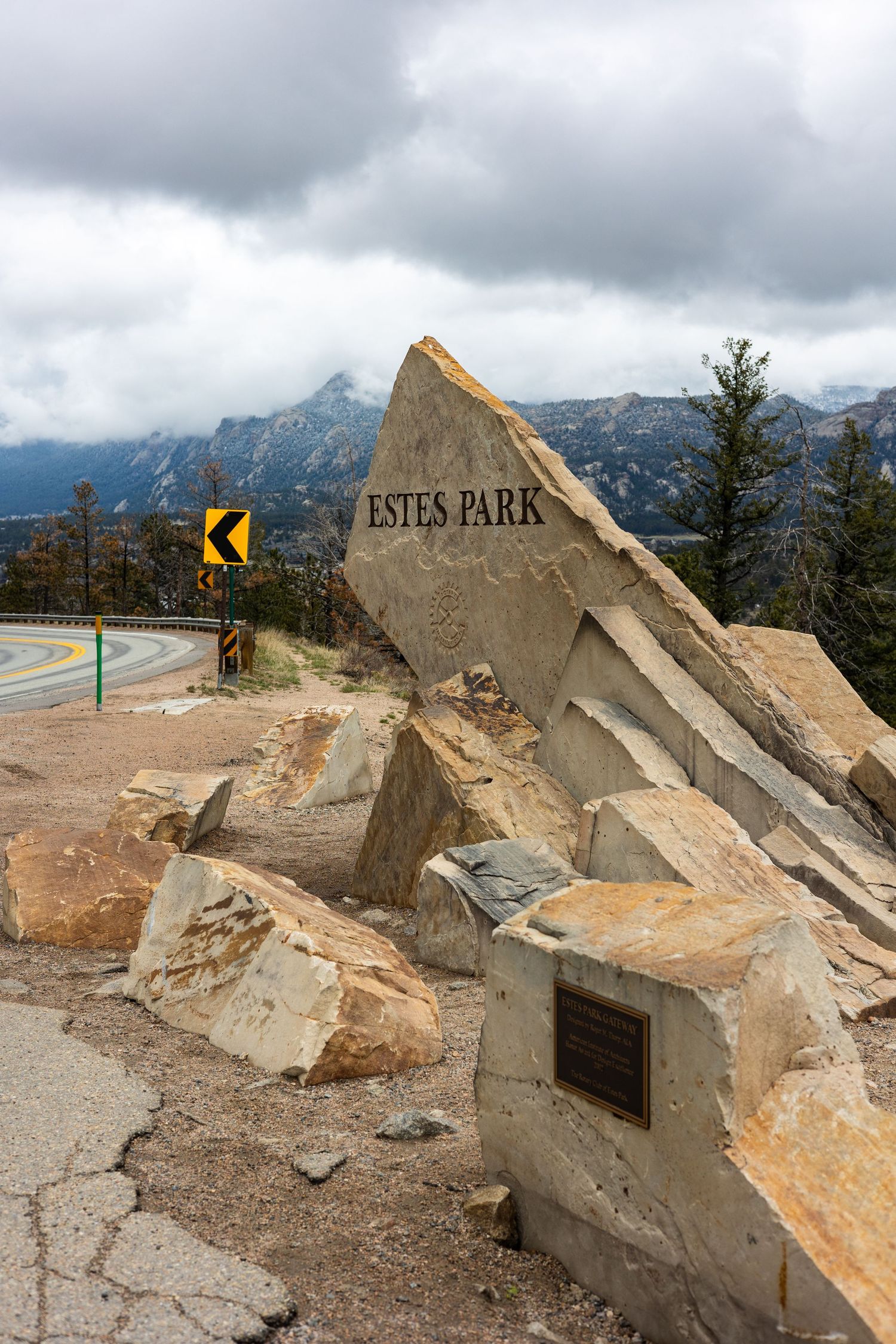 Entrance sign to Estes Park with mountain peaks and cloudy sky in background.