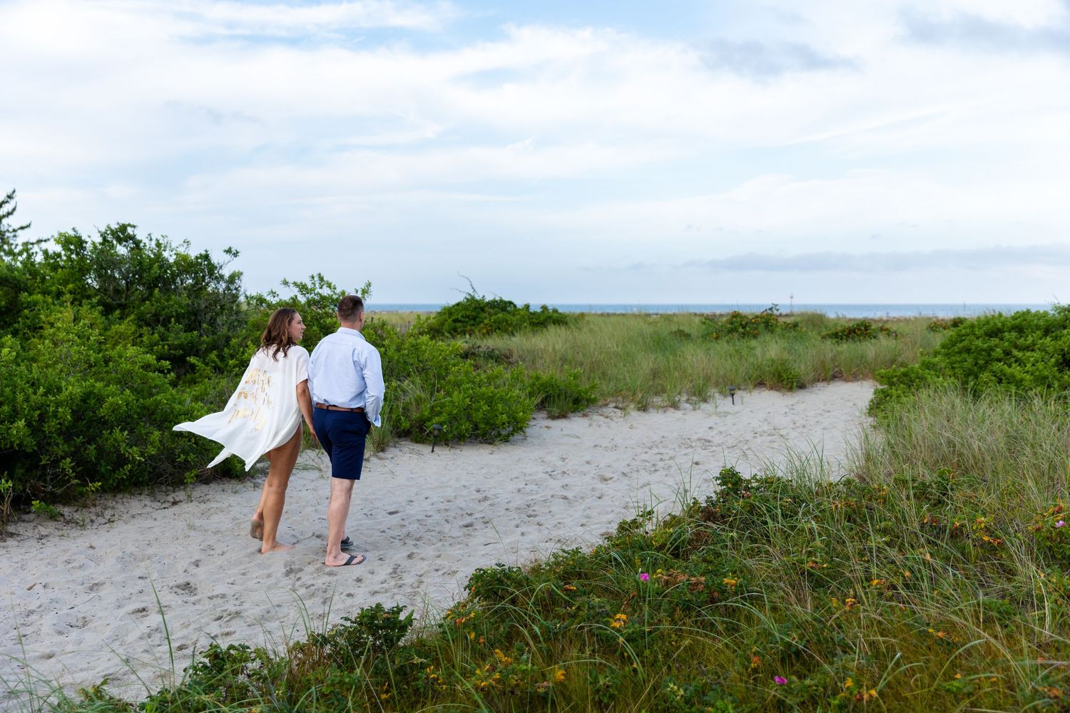 Two people walk along a sandy beach path surrounded by green coastal vegetation on a cloudy summer day.