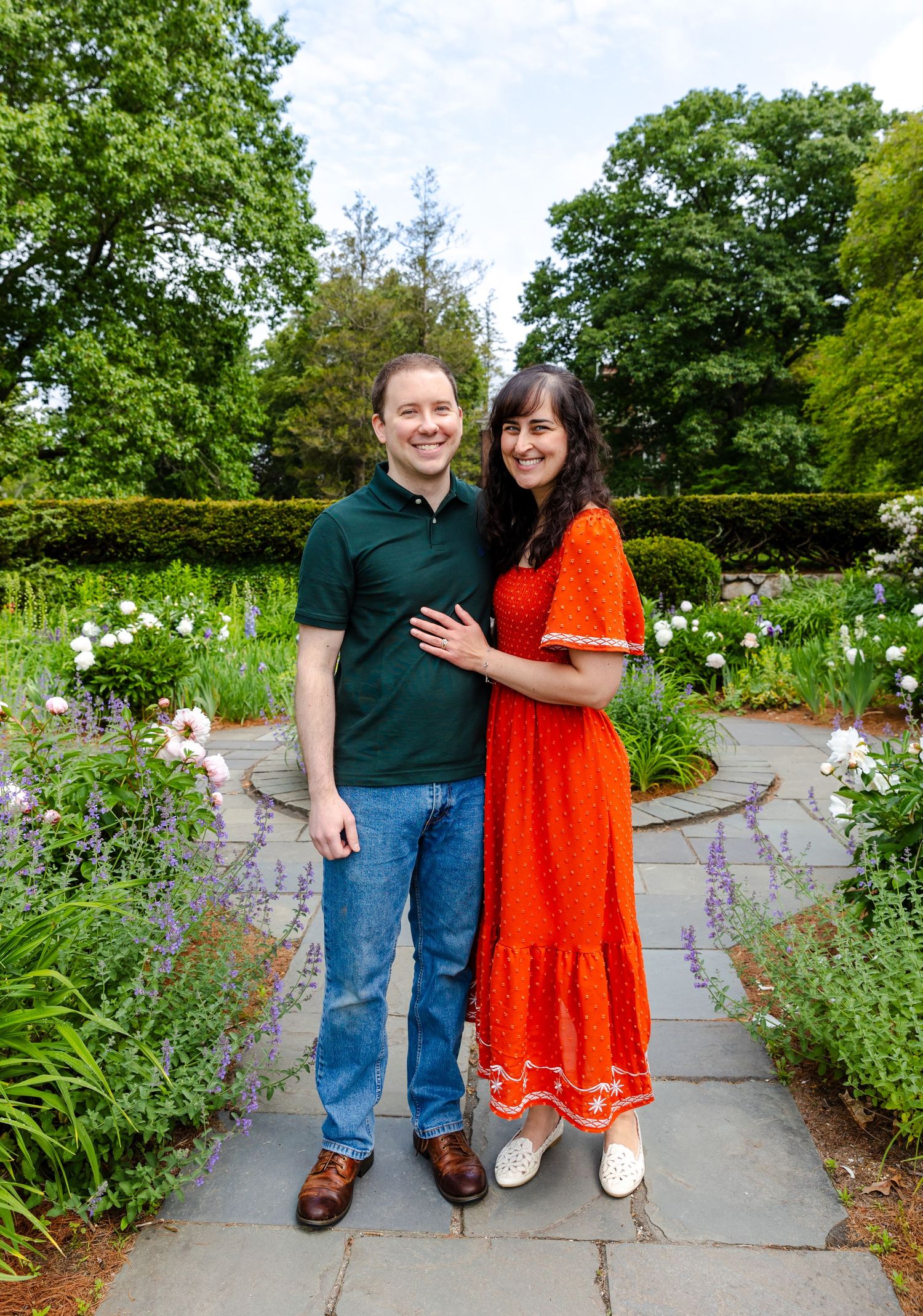 Two people smiling and posing together on a stone path surrounded by a lush garden with flowers and greenery.