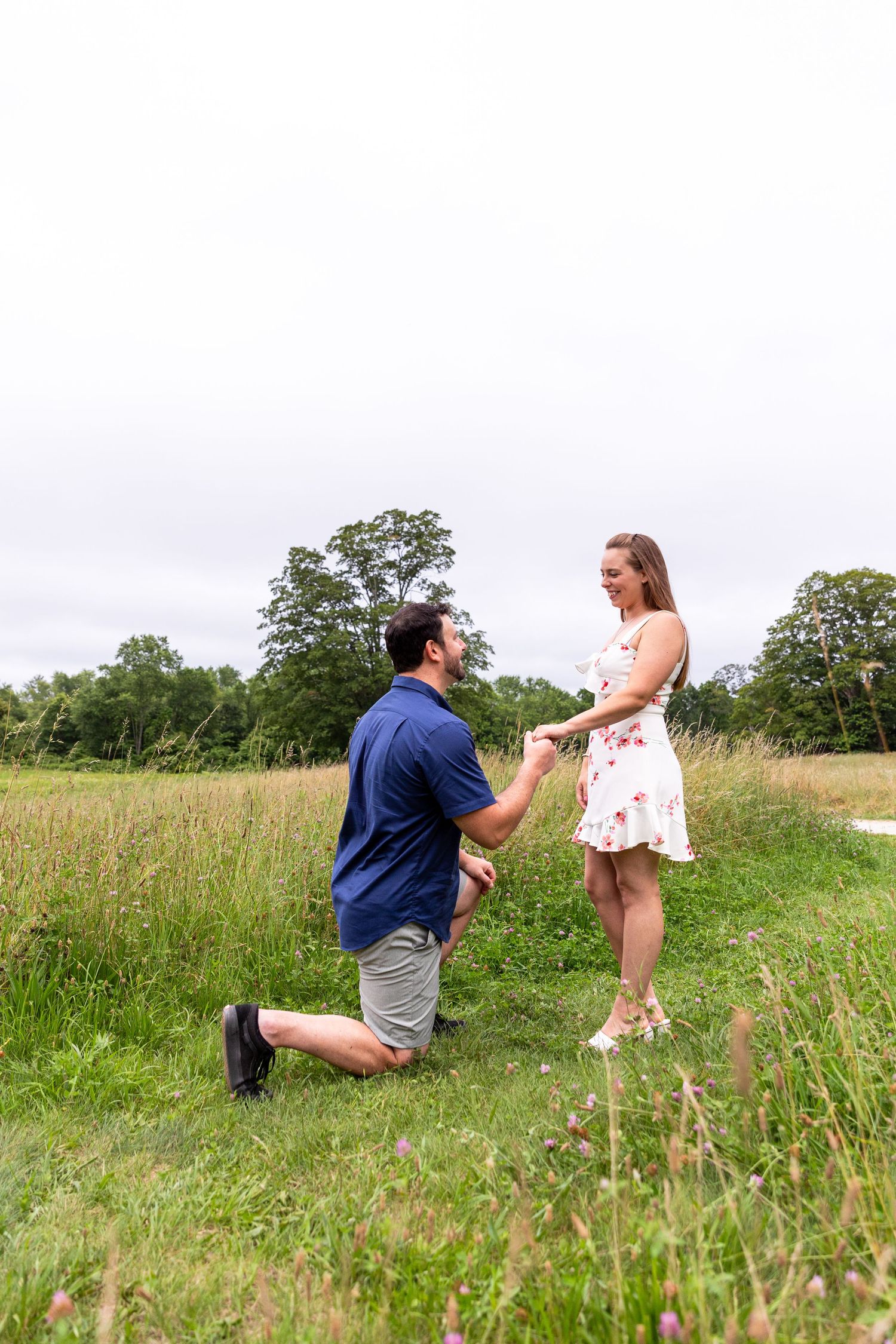 A romantic proposal moment captured in a summer meadow with tall grass and trees in the background.