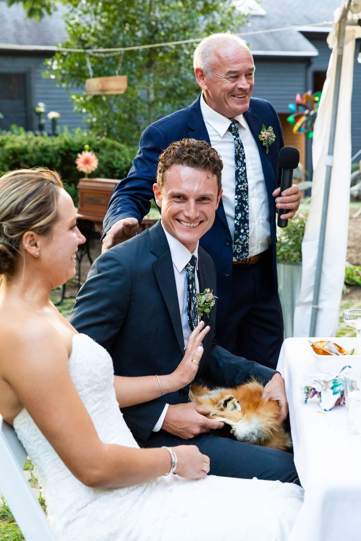 A wedding couple shares a joyful moment with a small dog at an outdoor reception.