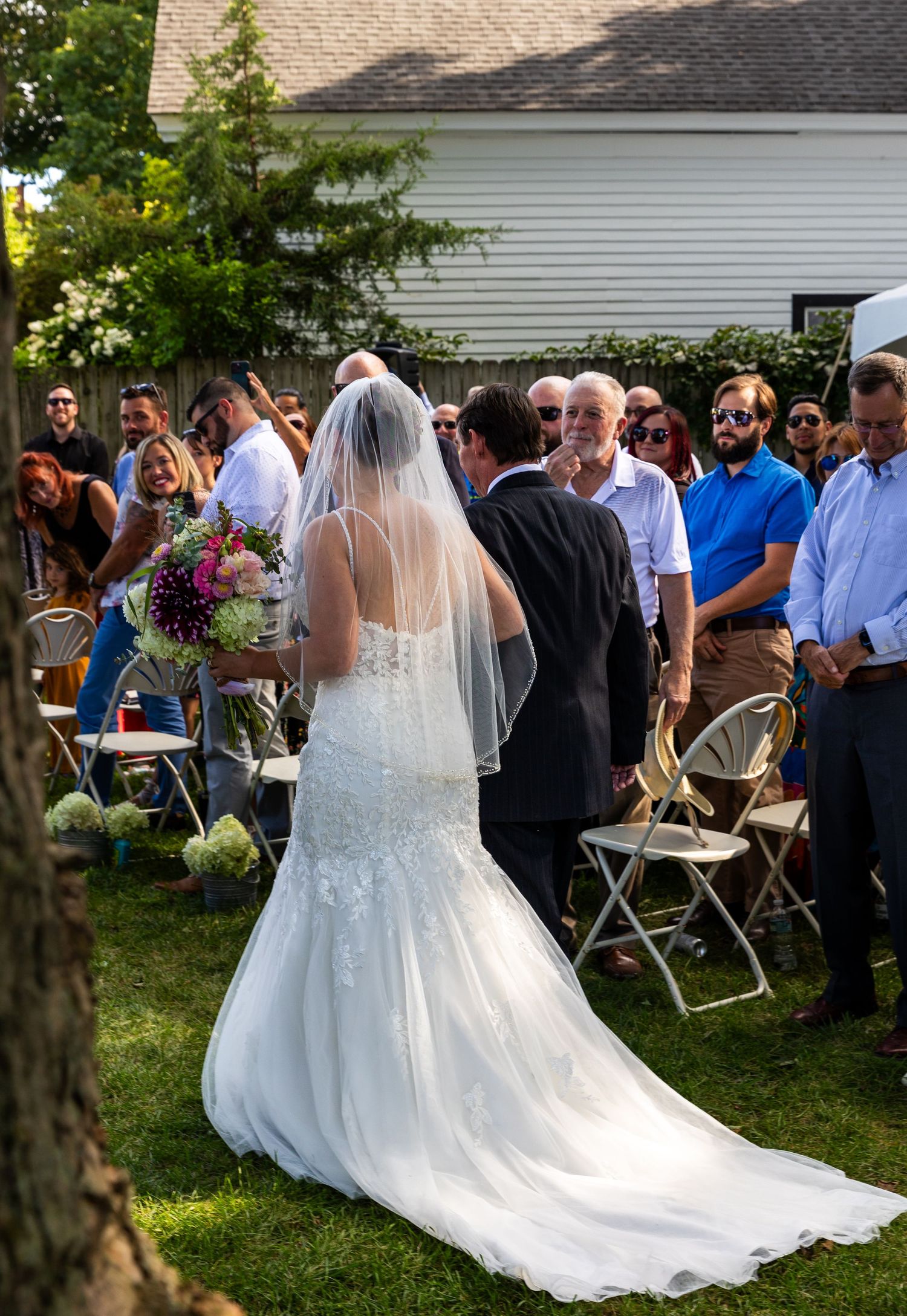Outdoor wedding reception scene showing backs of bride in flowing white gown and guests on a sunny lawn.
