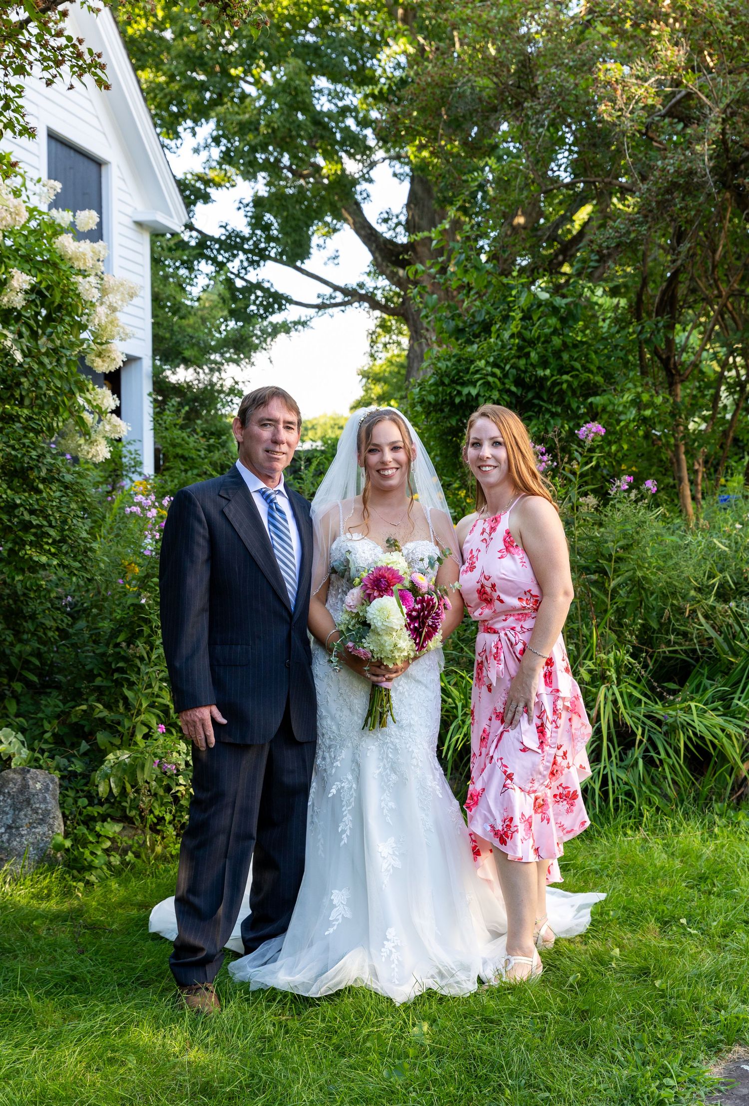 A wedding party poses for outdoor photos in a lush green garden setting with trees in the background.