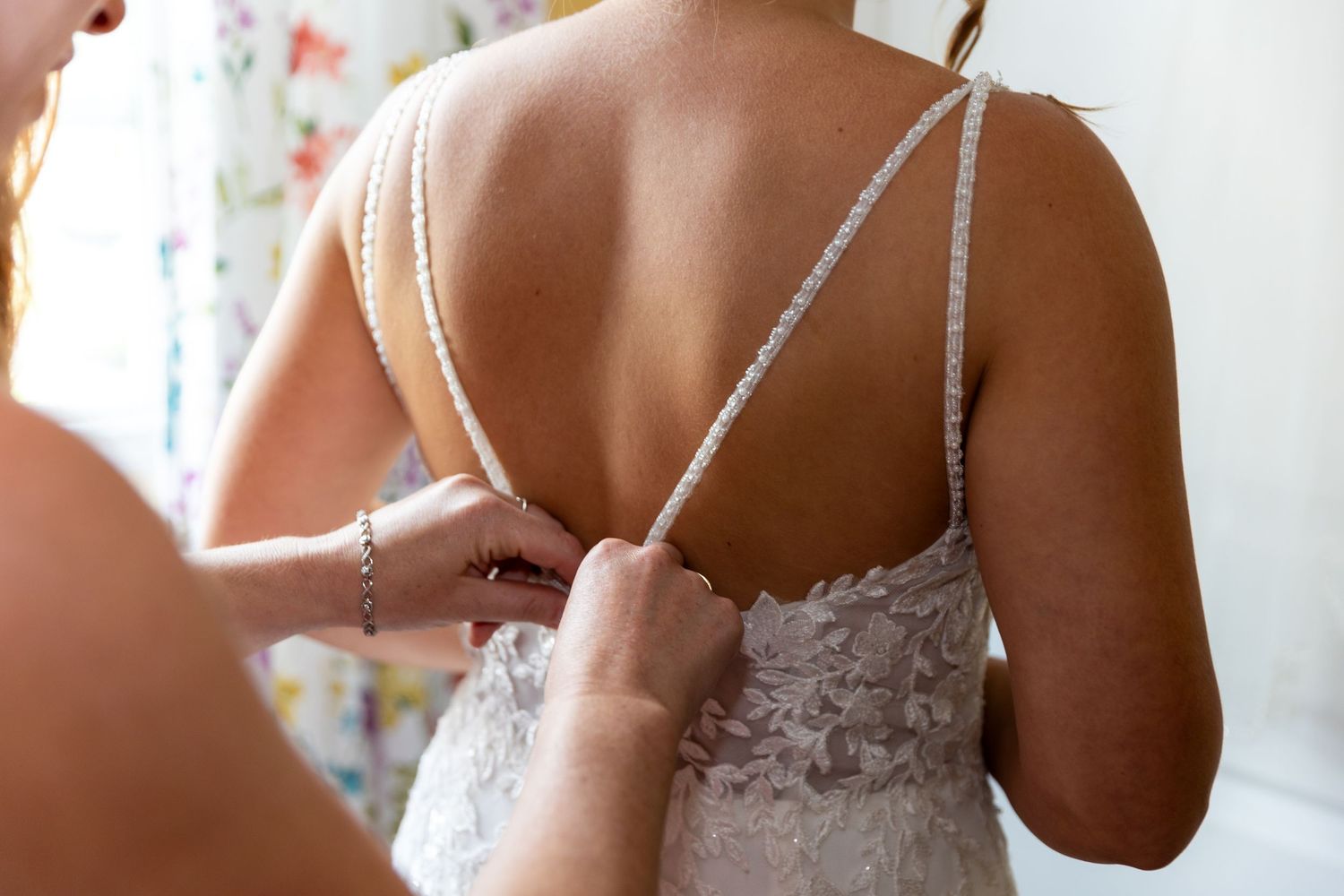 Someone helps fasten the delicate straps of a white lace wedding dress on a bride's back.