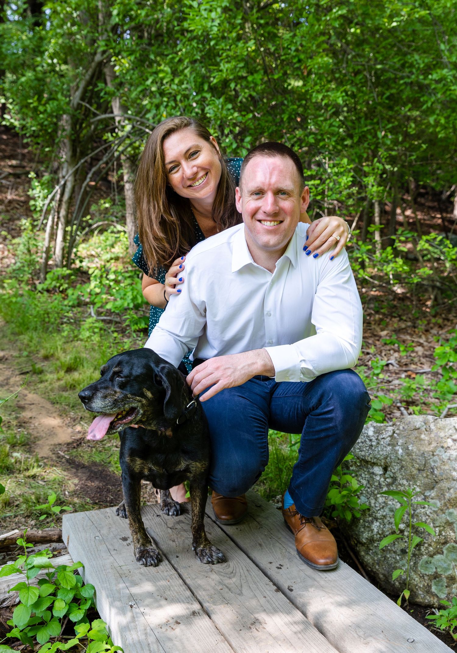 A black dog sits with someone on a wooden deck surrounded by lush green foliage in an outdoor setting.