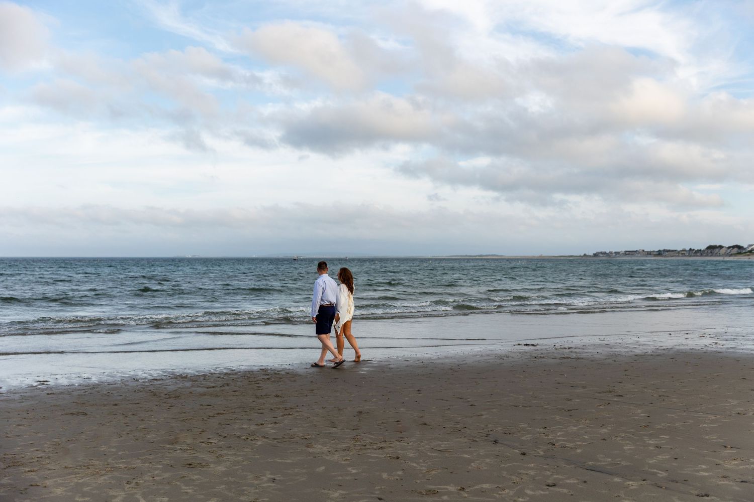 Several beachgoers enjoy a cloudy day along the shoreline as waves crash against the sandy coast at low tide.