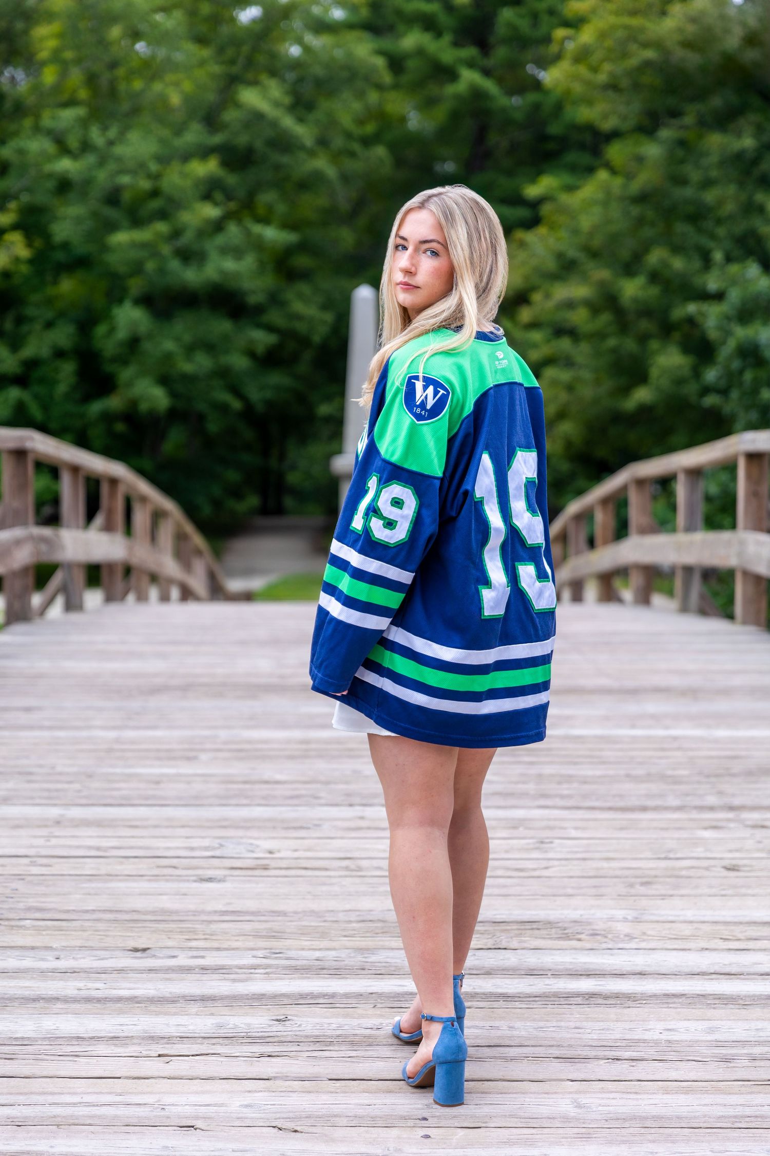 A person wearing a blue and green hockey jersey poses on a wooden bridge.