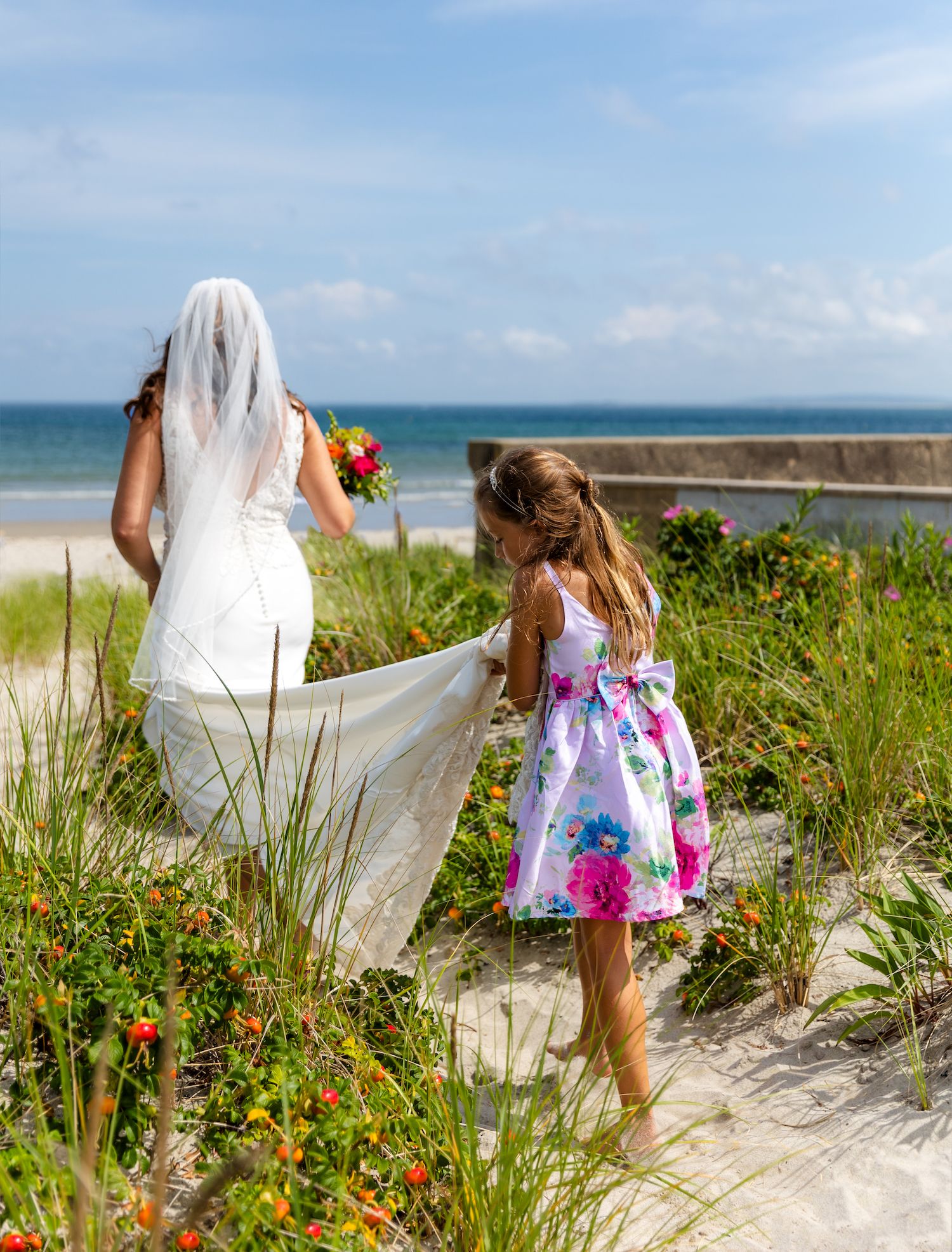 A bride and flower girl walk along a beach path with ocean views and wildflowers.