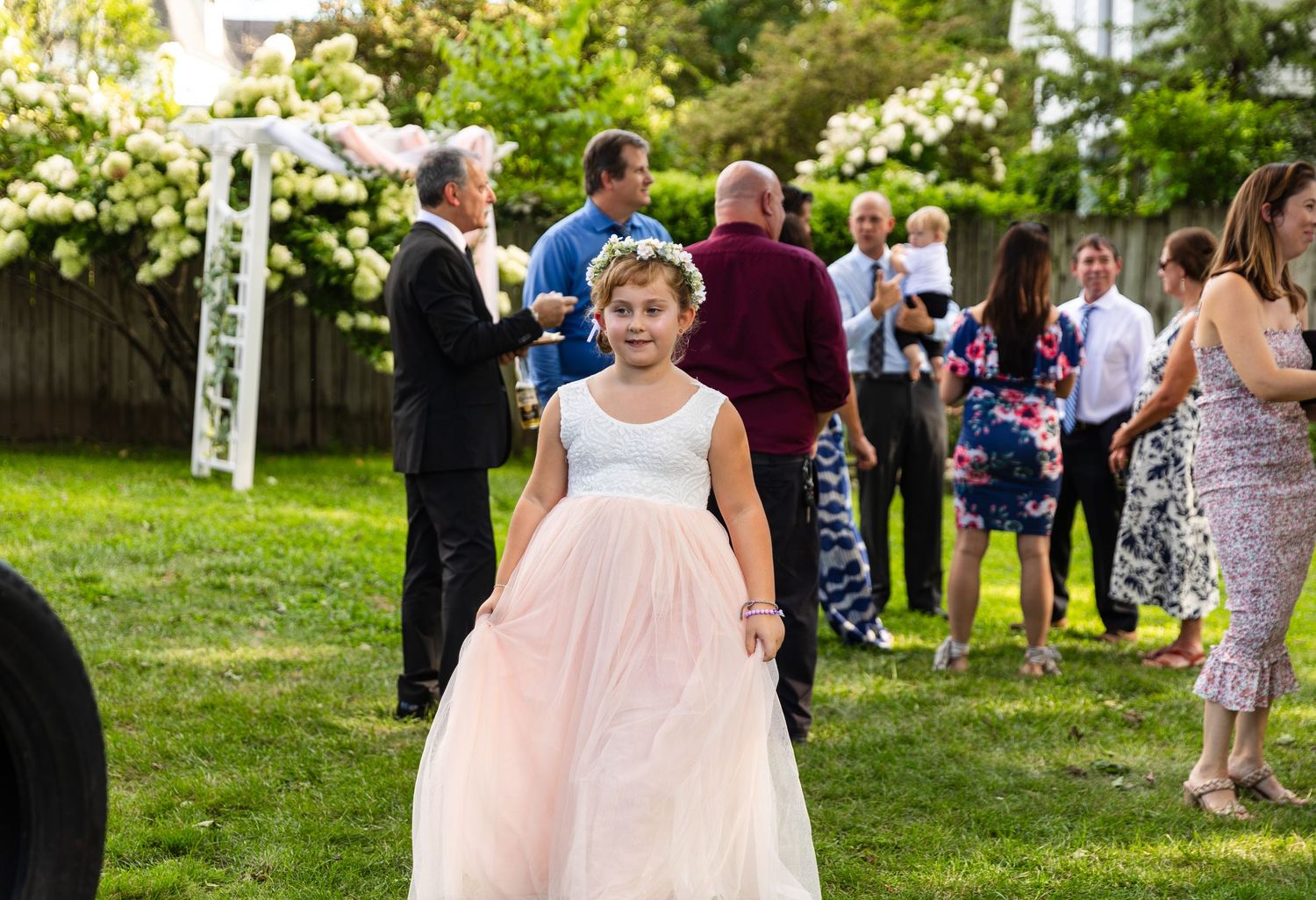 A person in a flowing pink ballgown walks across a sunny outdoor garden party setting with guests mingling in background.