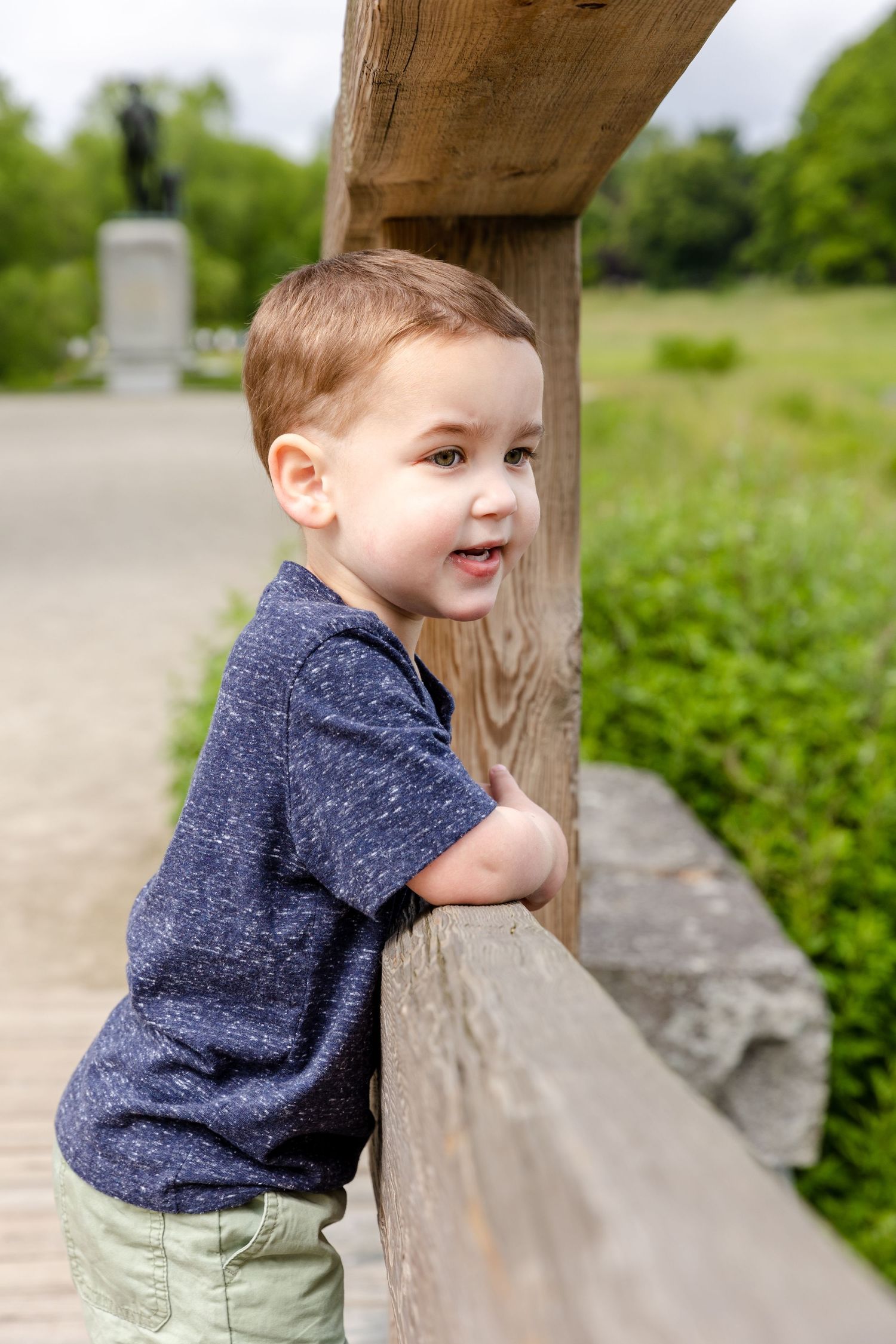 Young child in a navy blue shirt and khaki shorts leans on a wooden railing in an outdoor park setting.