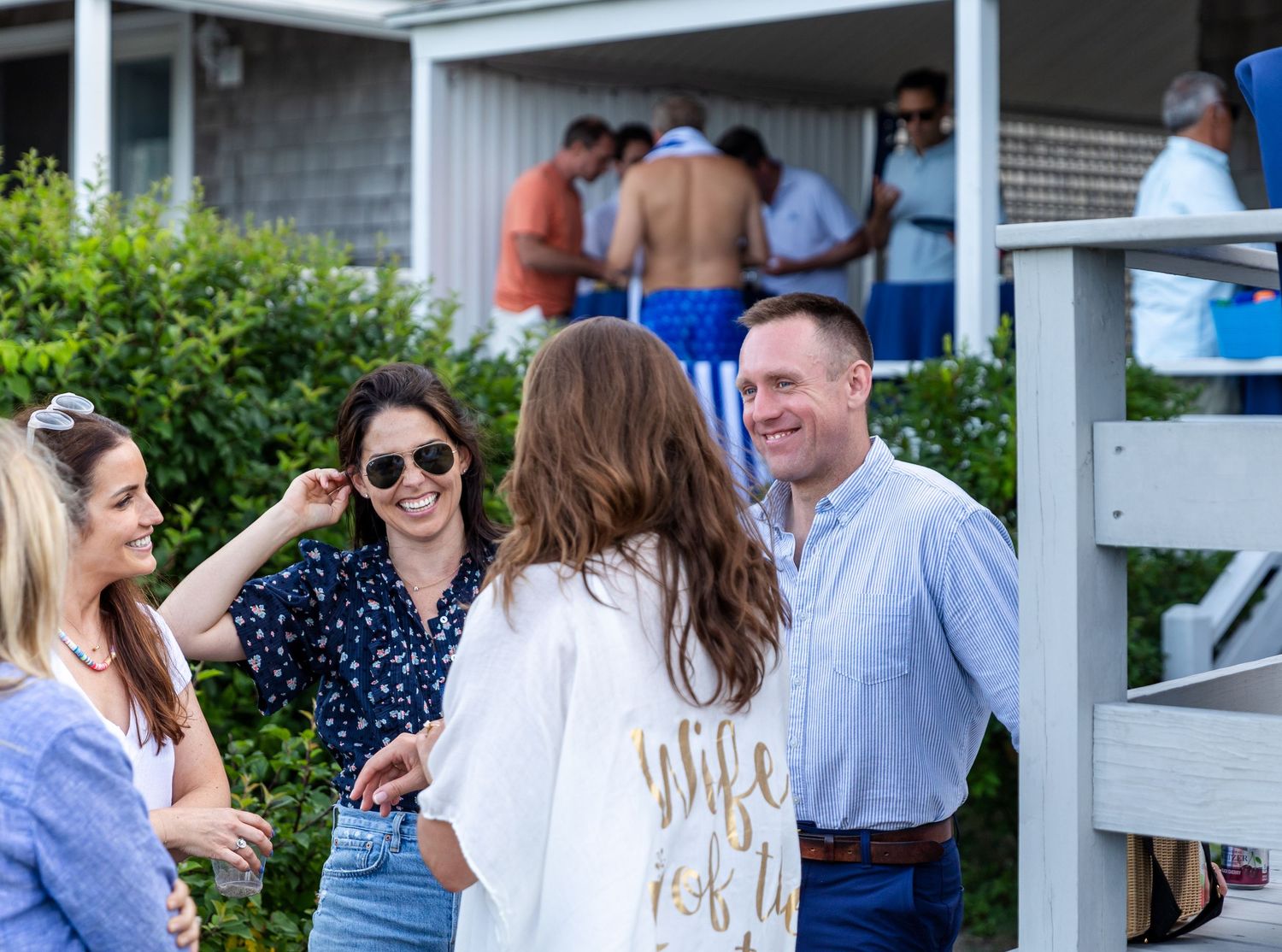 Group of people socializing and enjoying themselves at an outdoor gathering near bushes and a building.