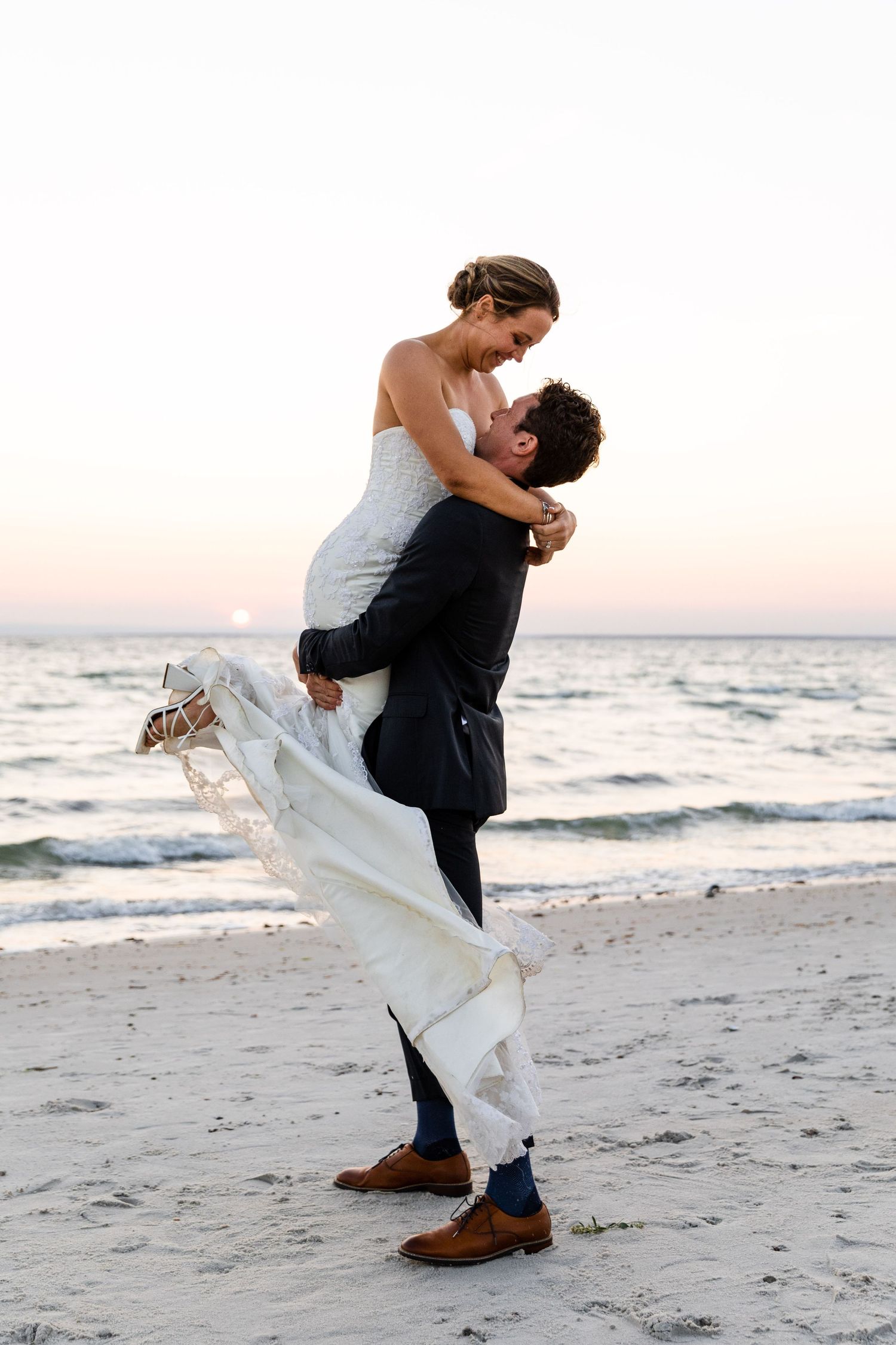 A couple shares a romantic dip kiss moment on a sandy beach at sunset with ocean waves in the background.