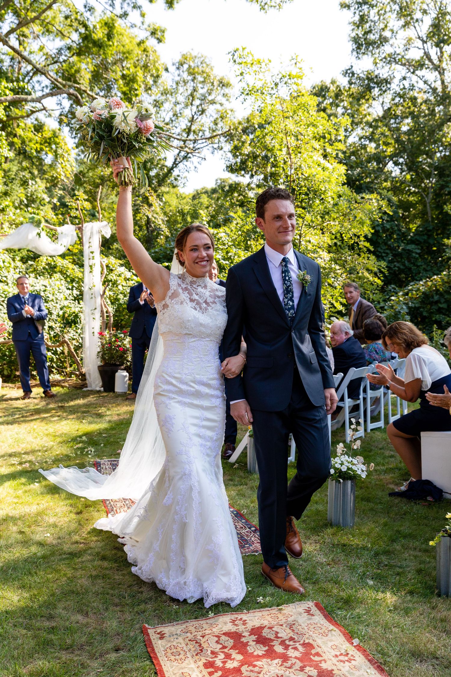 A couple celebrates their wedding ceremony on a sunny lawn with decorative floral arch and vintage rug.