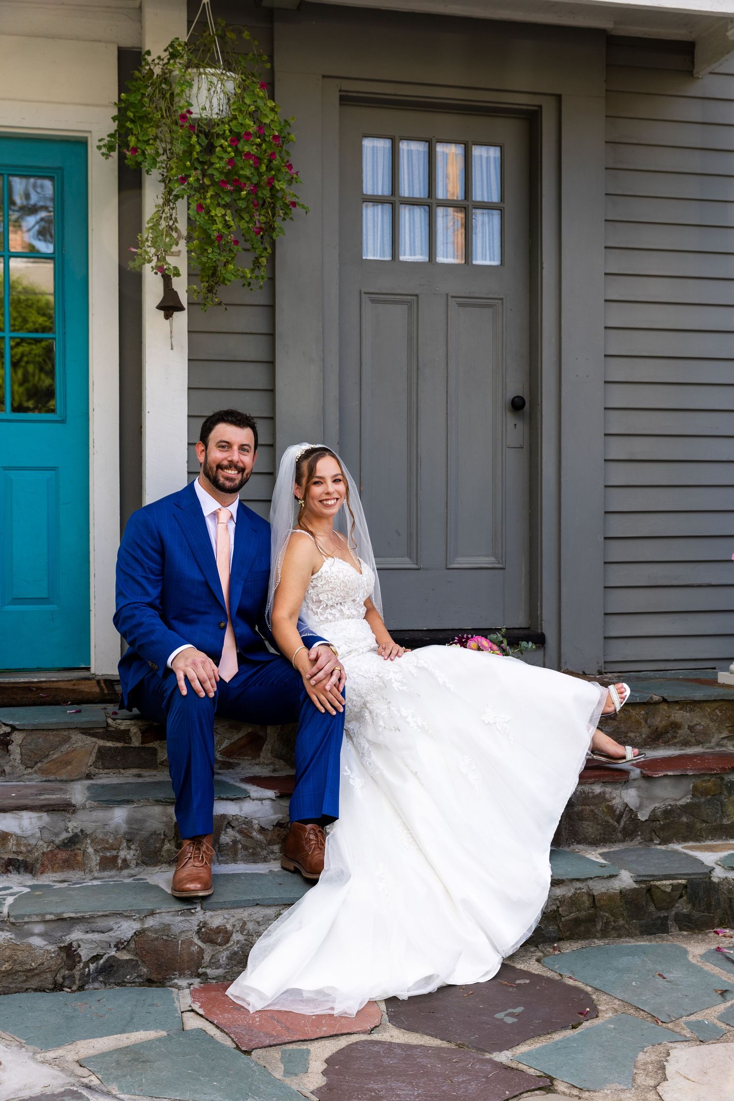 A couple in formal wedding attire poses playfully on gray stone steps in front of a house with teal blue doors.